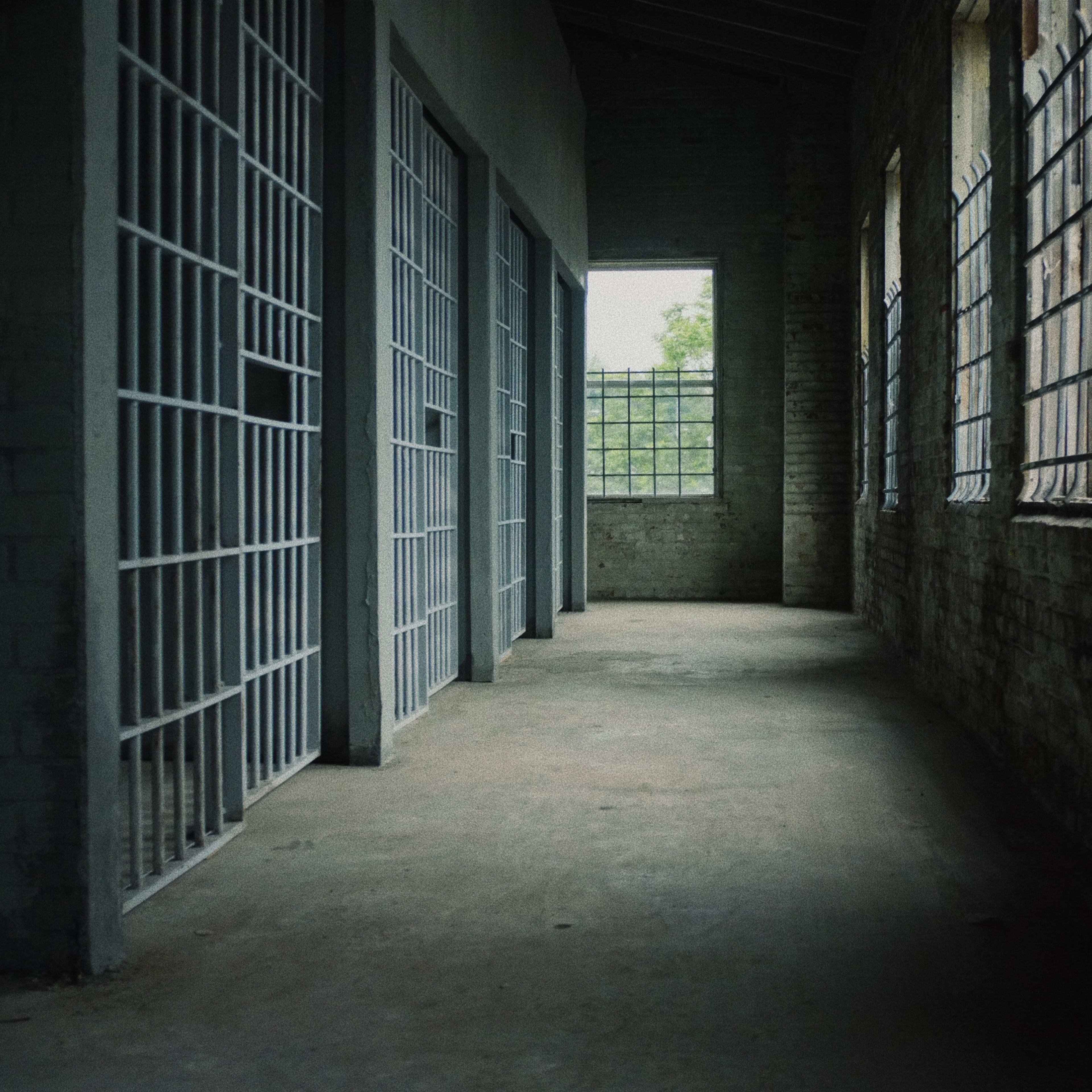 A dimly lit hallway in an old building with empty jail cells on either side and windows allowing natural light to enter.