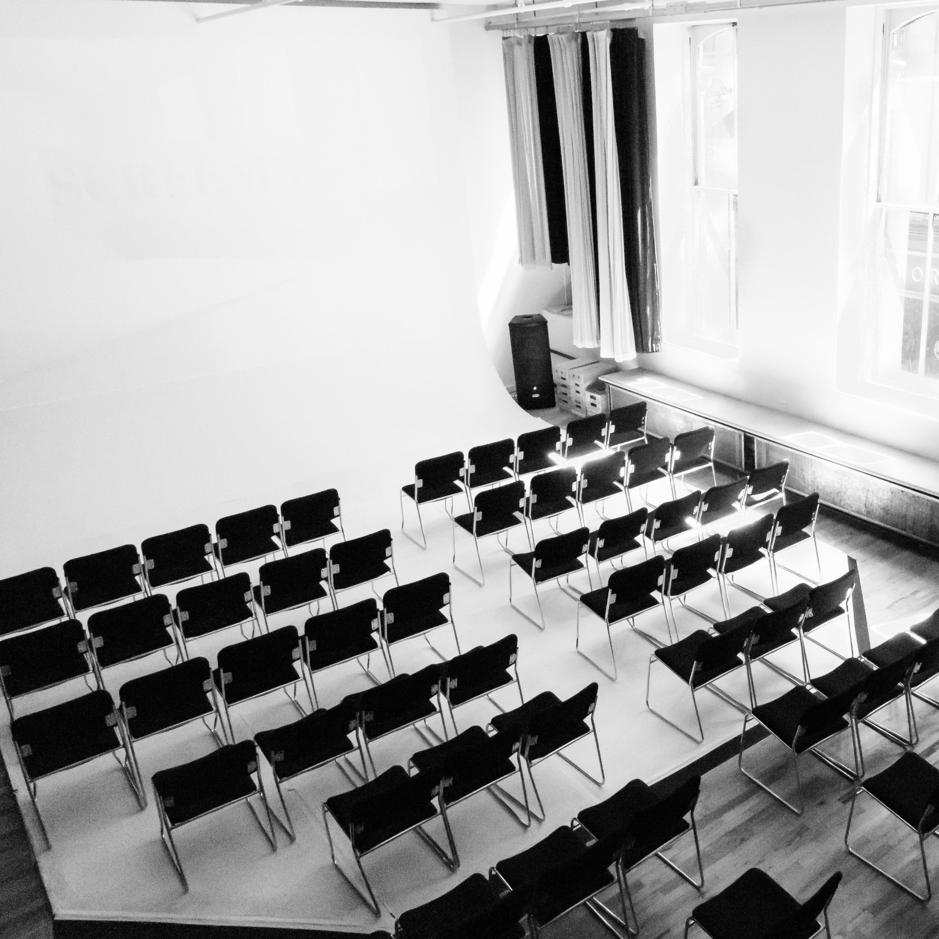 A row of black chairs is arranged in a semi-circle facing a blank wall in a well-lit room with large windows.
