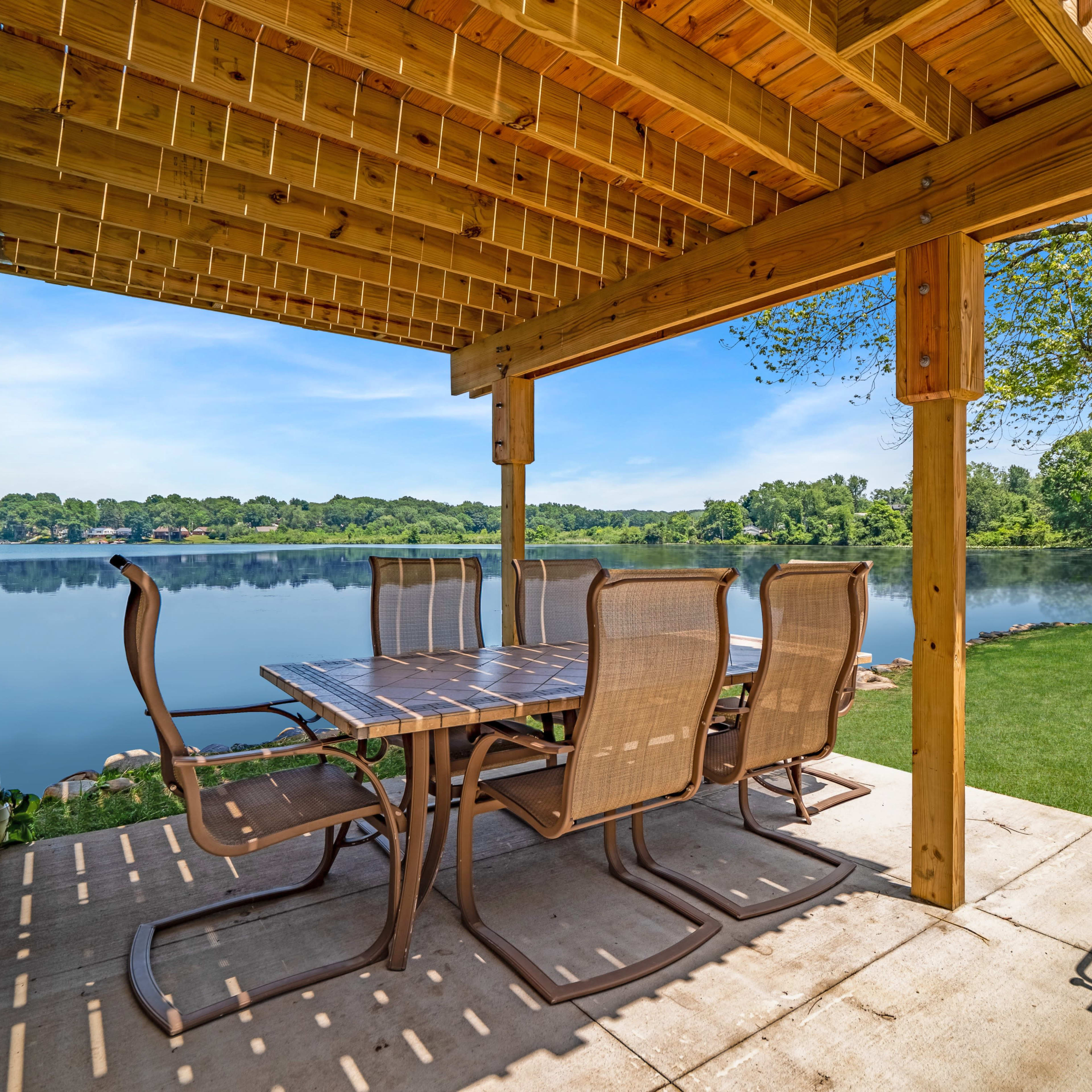 A patio area with a large table and chairs overlooks a calm lake, framed by a wooden pergola and tree.