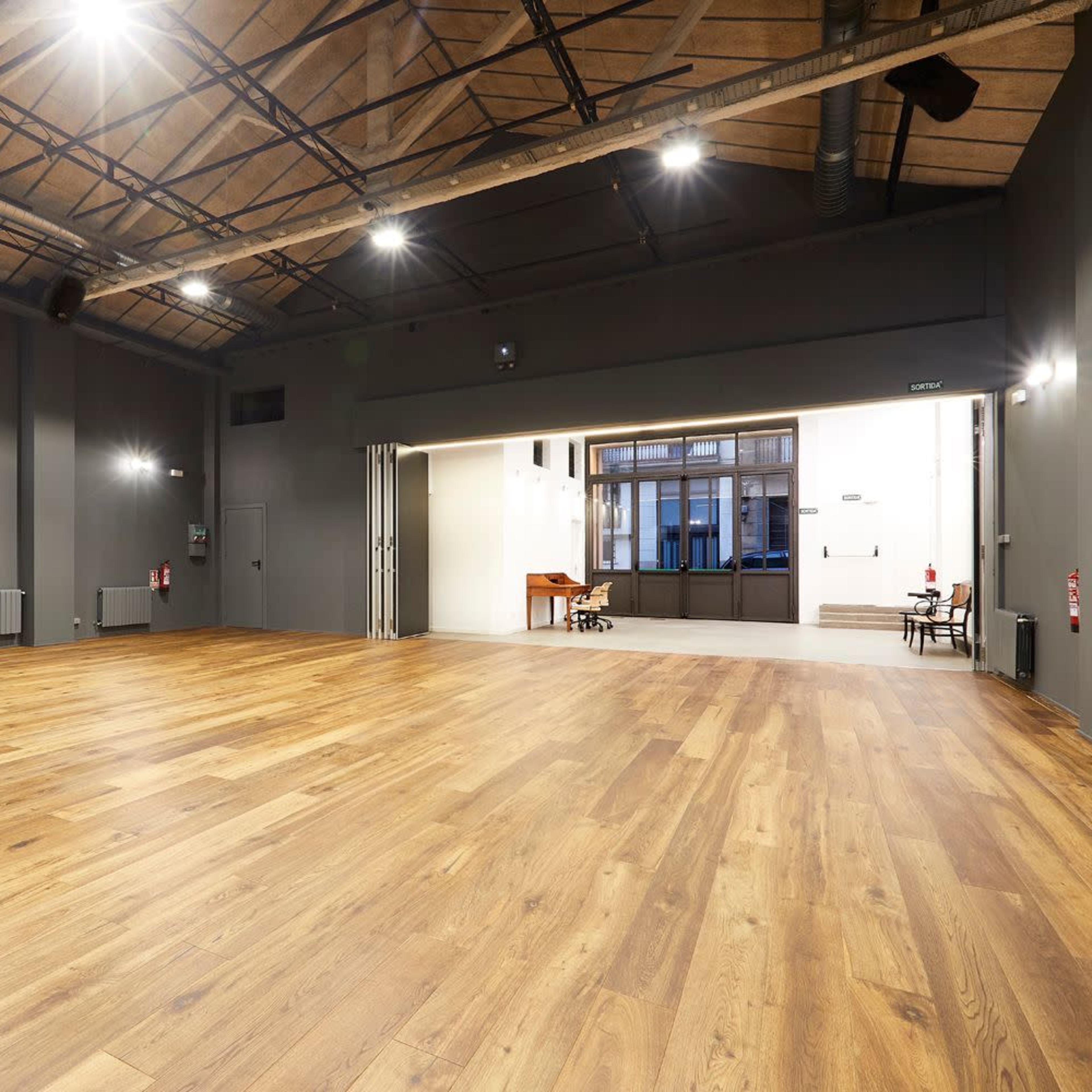 A spacious, empty hall with wooden flooring and high ceilings, featuring gray walls and soft lighting.