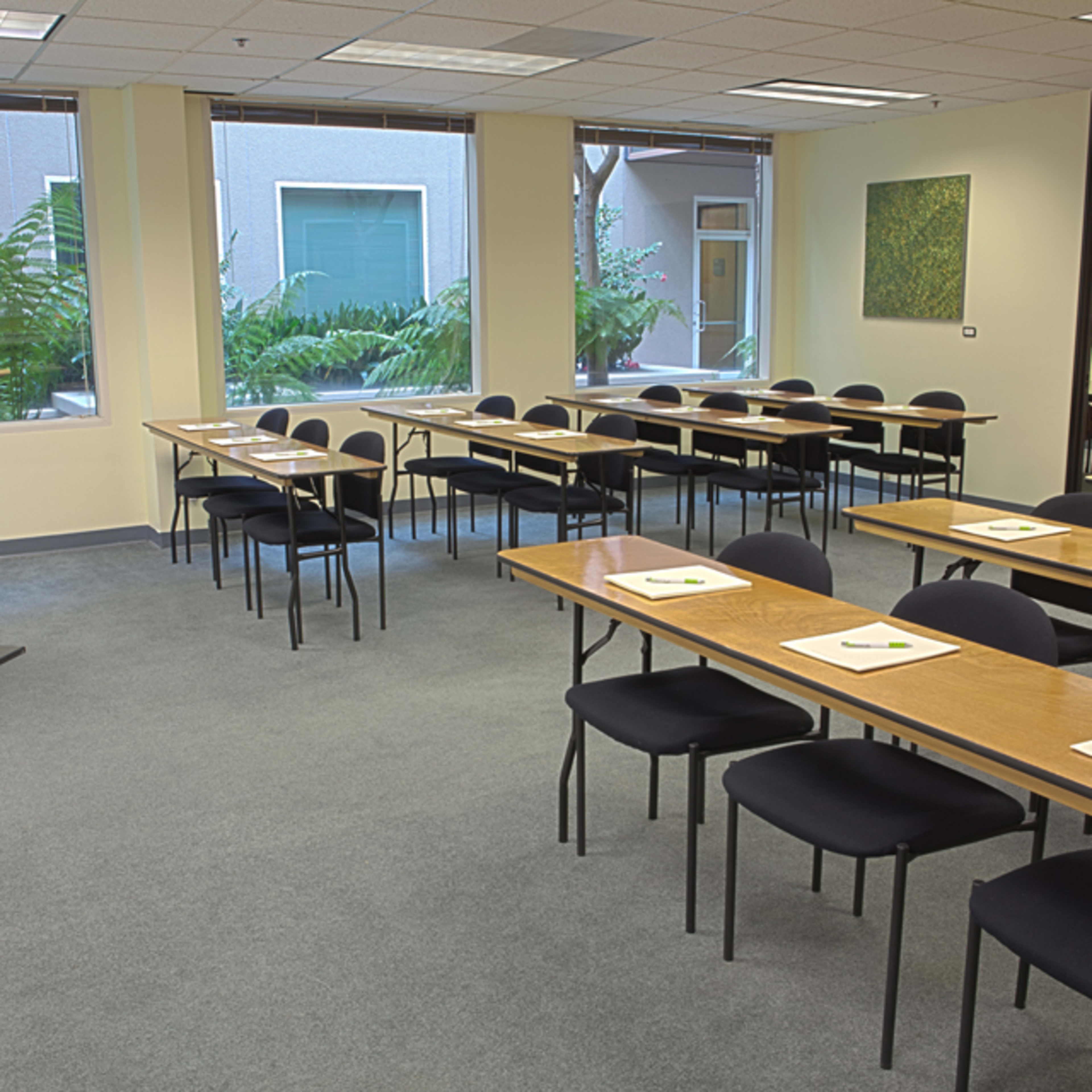 A conference room is set up with multiple tables and chairs, facing a podium, and features large windows overlooking a green courtyard.