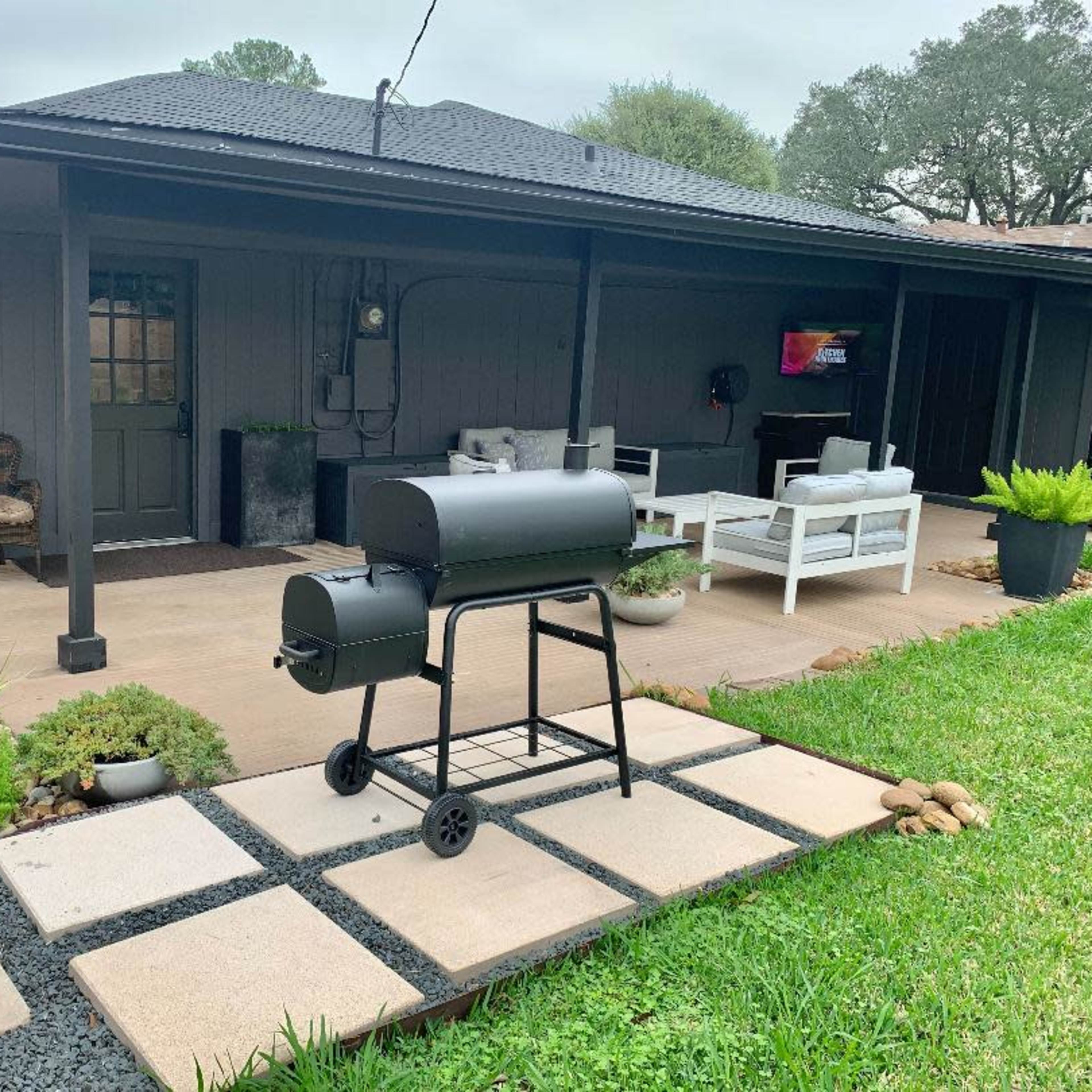 A backyard patio with a charcoal grill on a stone walkway, surrounded by greenery and outdoor furniture.