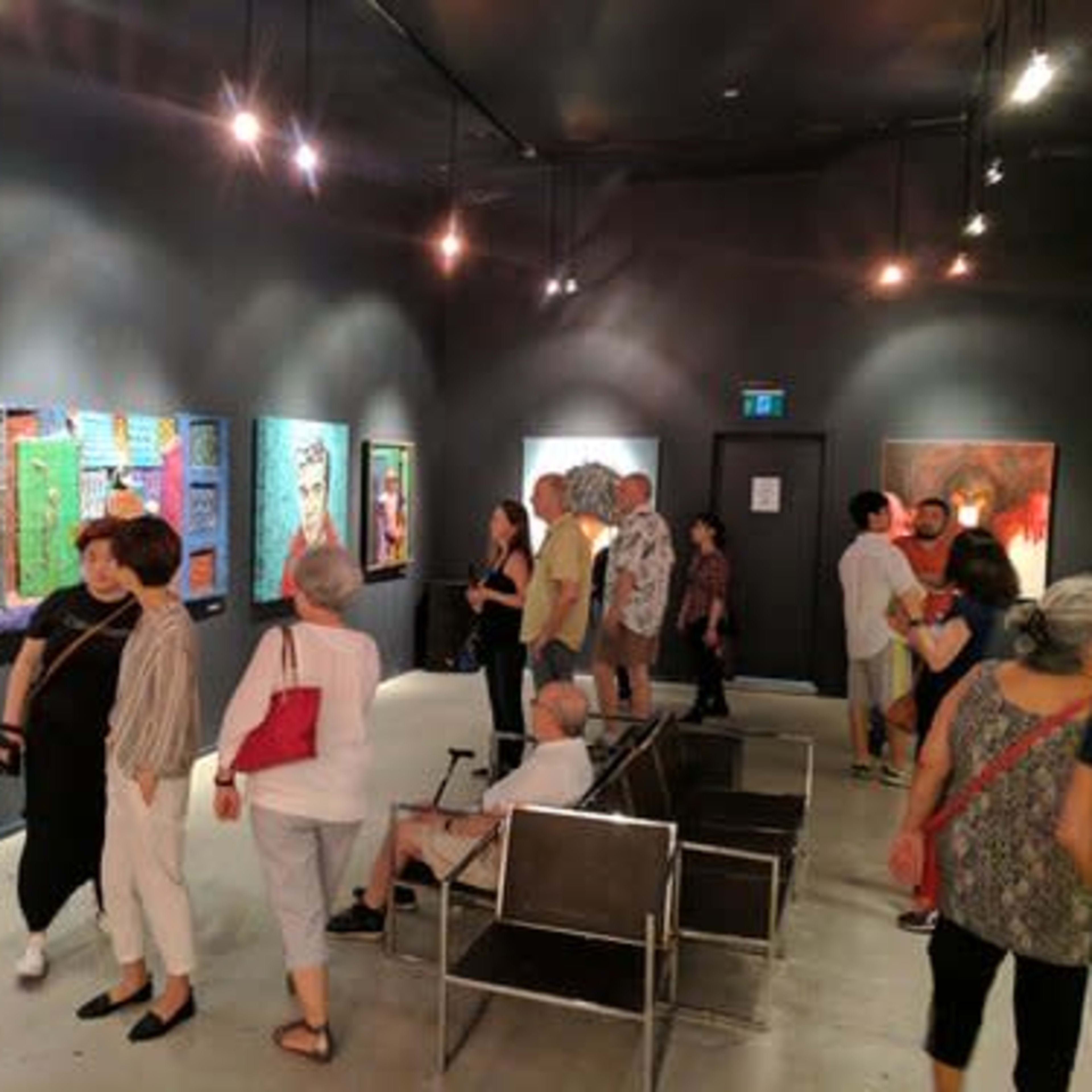A crowded art gallery features various colorful paintings on the walls while visitors chat and observe the exhibits.