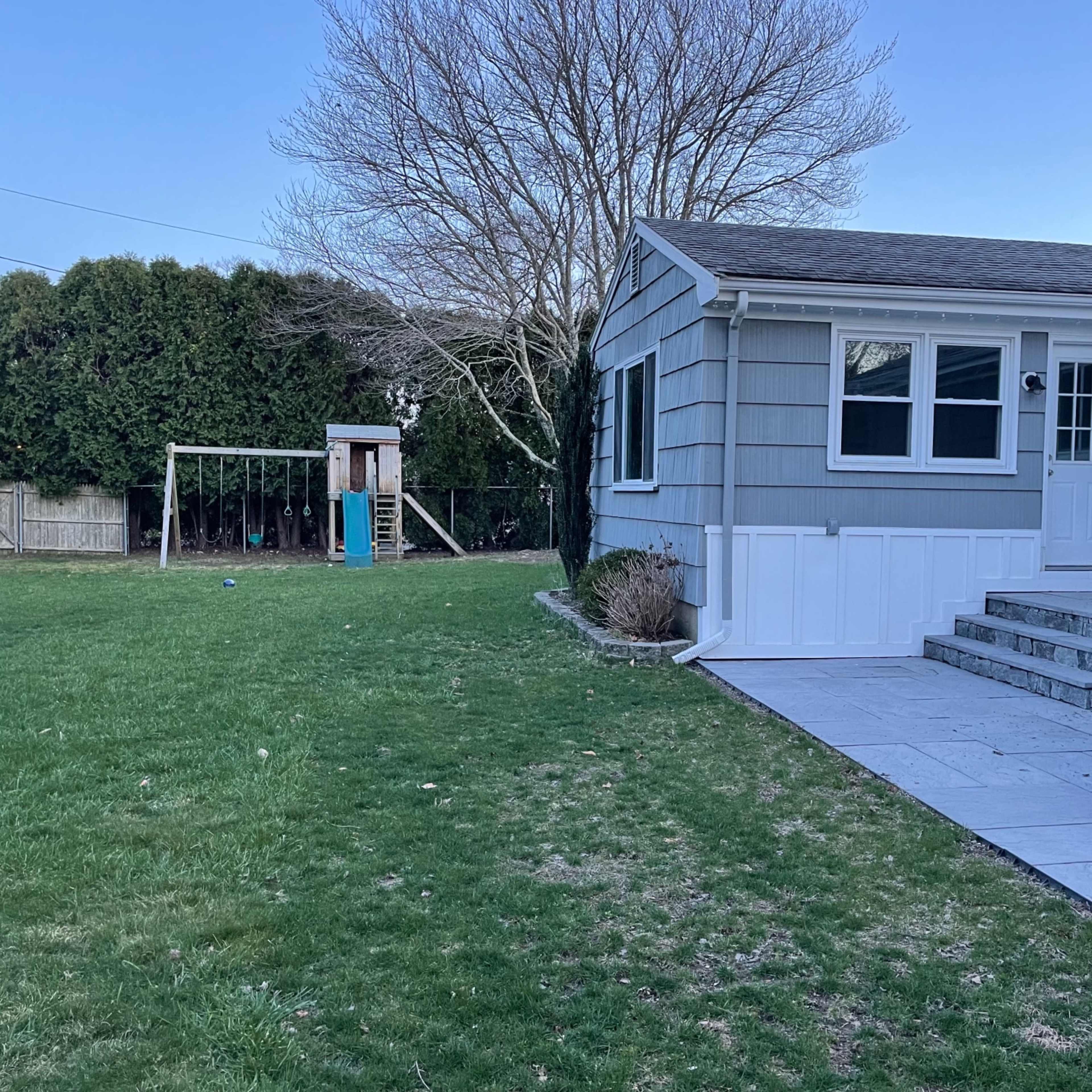 A backyard features a patio area with a table and chairs adjacent to a playset and a grassy lawn.
