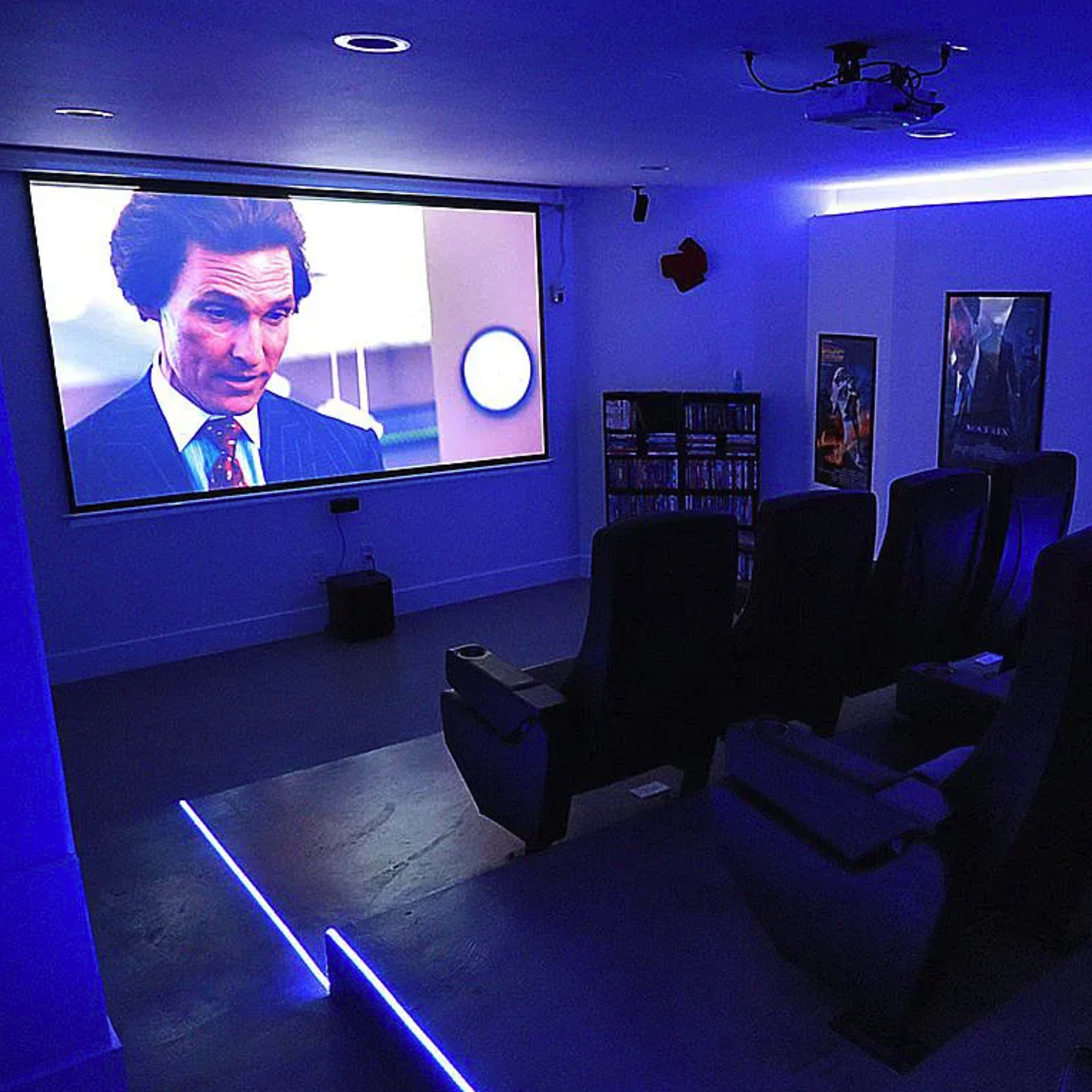 A home theater featuring a large projection screen displaying a movie scene, with several black reclining seats arranged in front and movie posters on the walls.