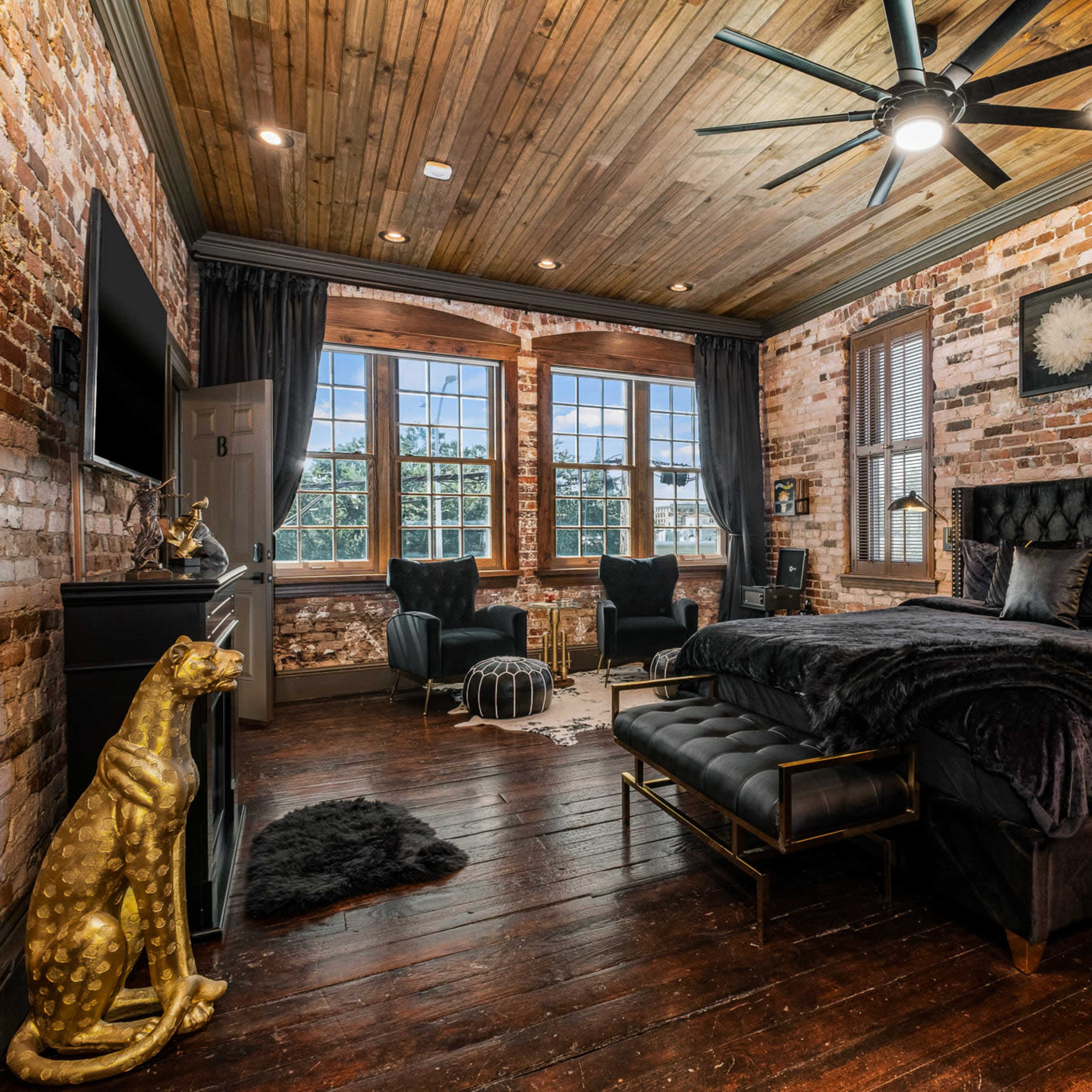 A cozy bedroom featuring exposed brick walls, a wooden ceiling, a king-size bed with dark bedding, and two armchairs near large windows.