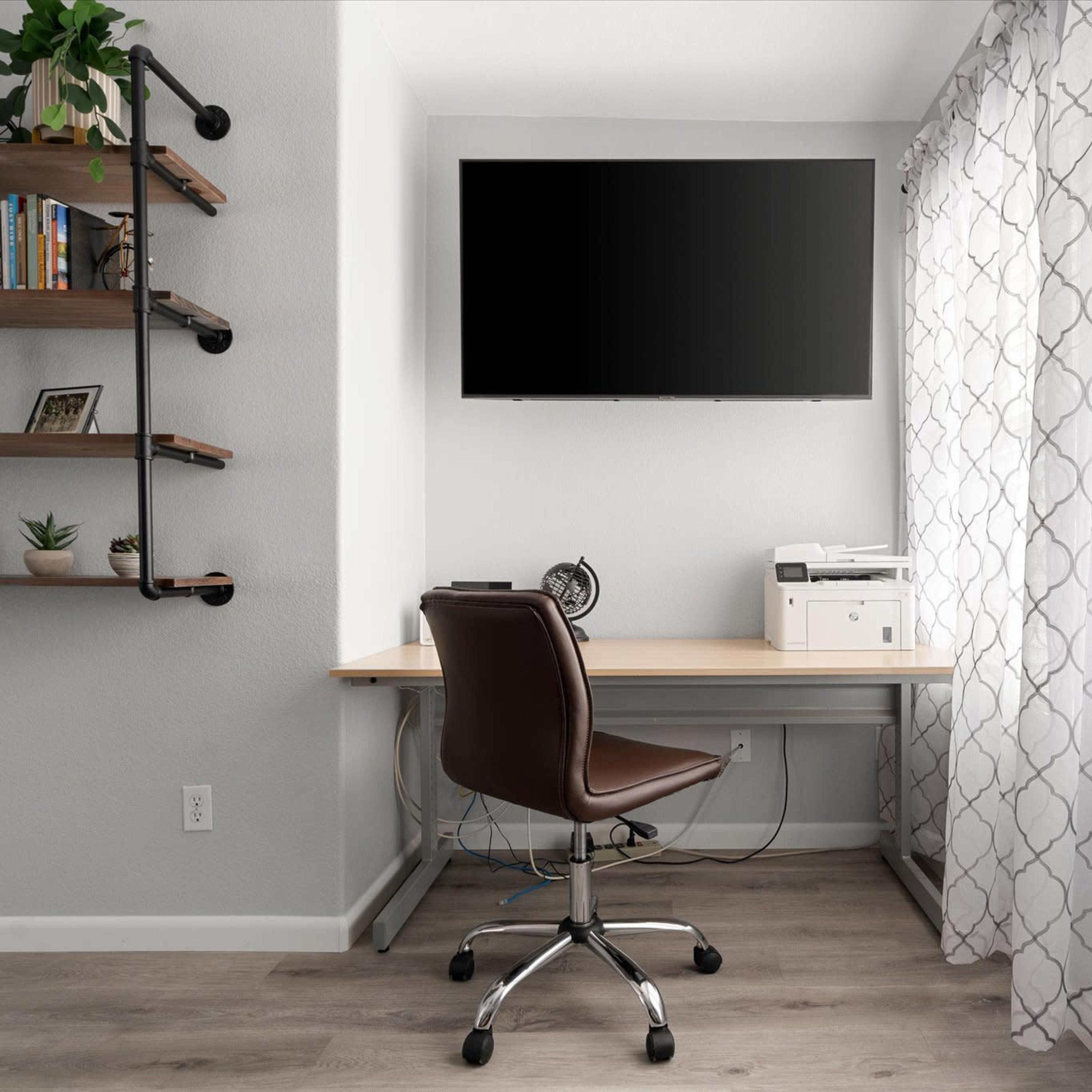 A modern home office setup with a wooden desk, a swivel chair, a printer, a wall-mounted TV, and a shelf filled with plants and books.