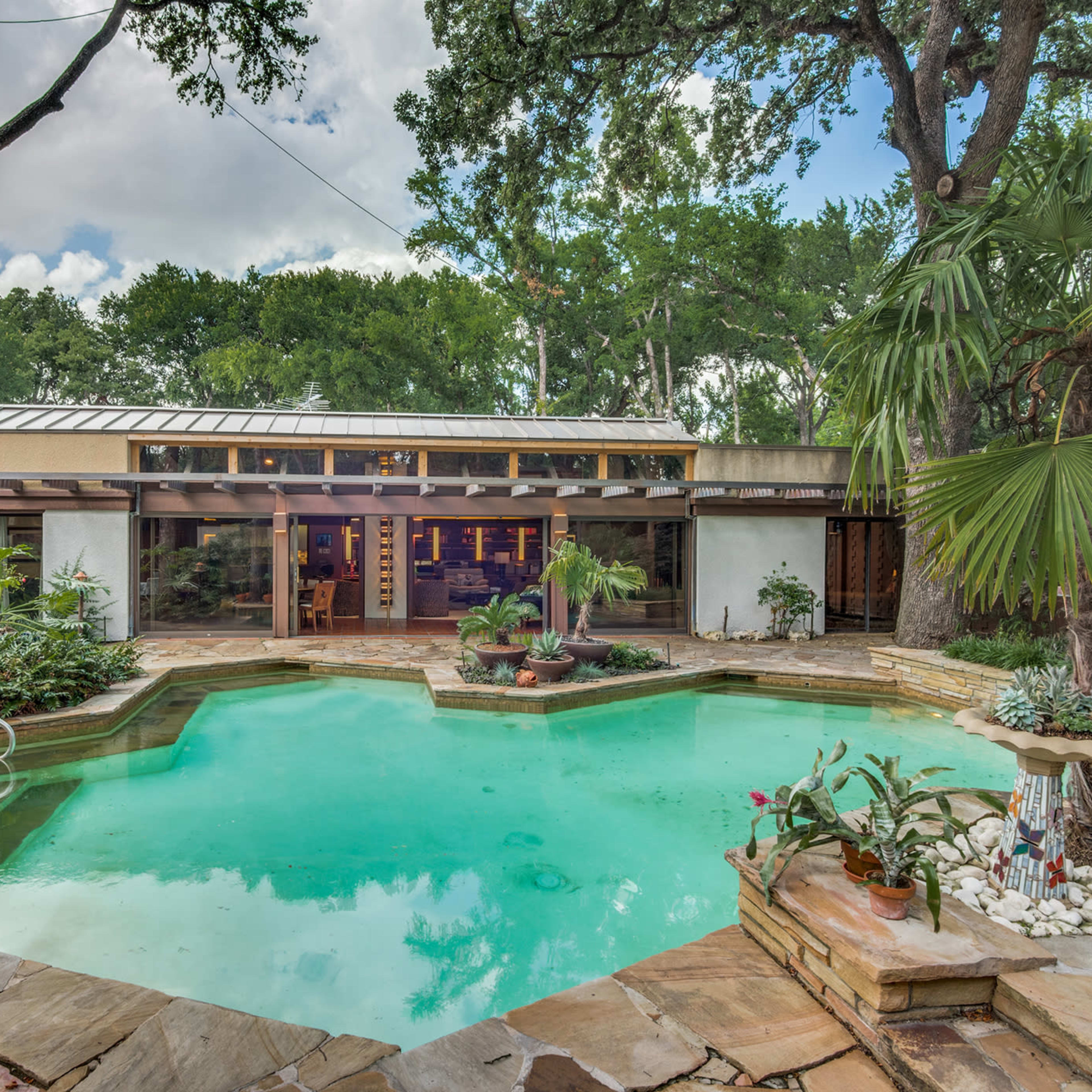 A backyard with a green pool surrounded by stone decking, tropical plants, and a house in the background with large windows.