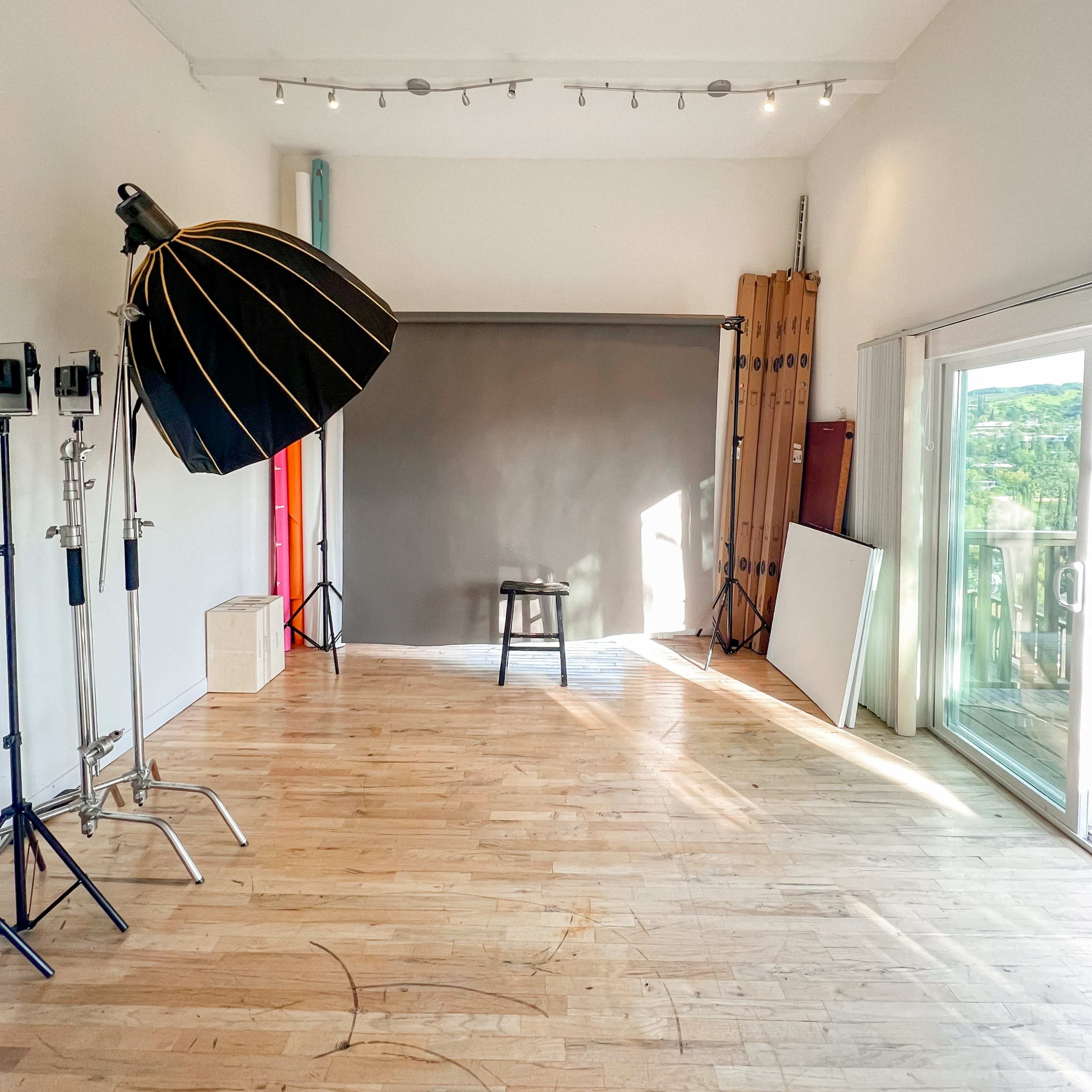 A photography studio with a gray backdrop, a stool, and various lighting equipment arranged against a wooden floor and large windows.