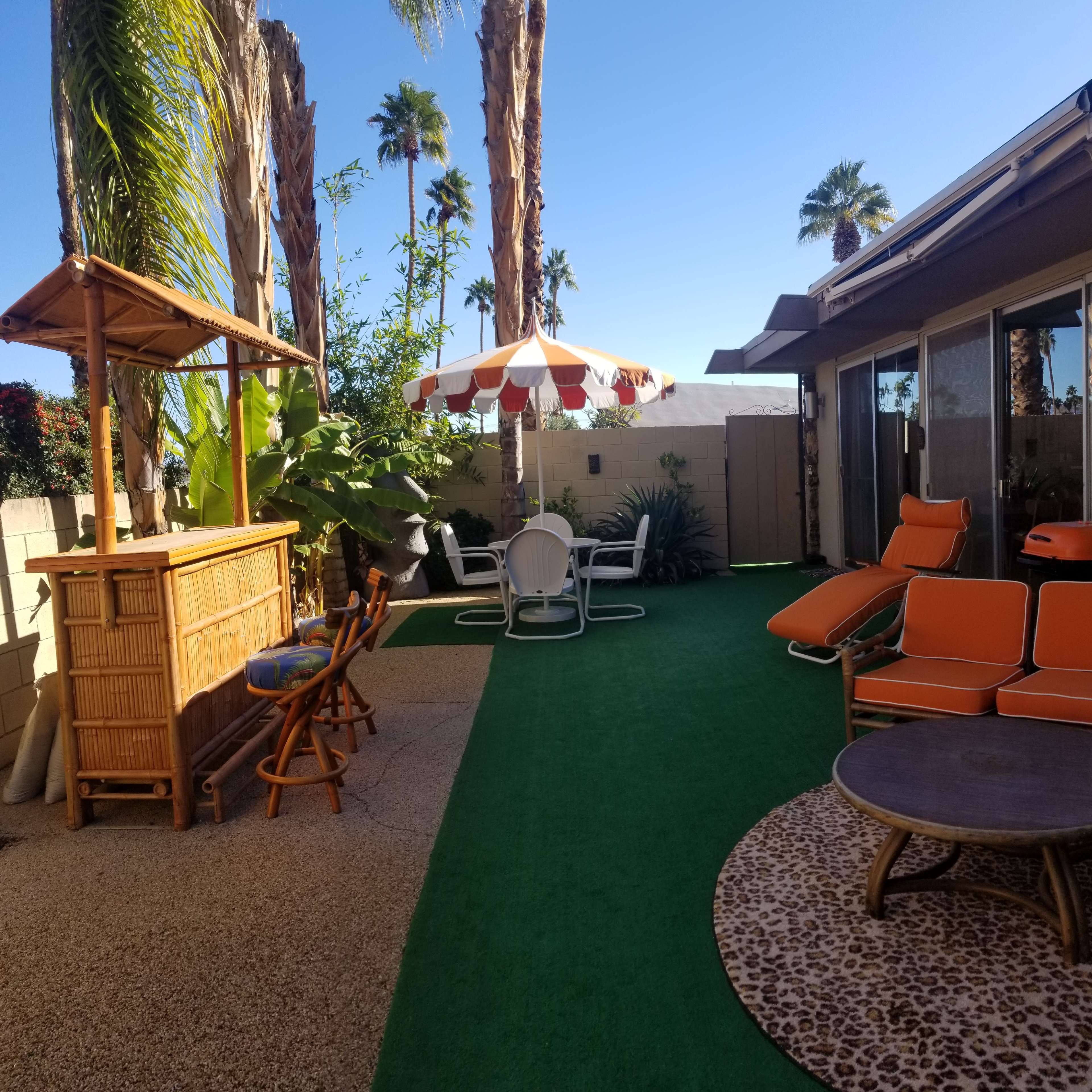 A backyard patio features a bamboo bar, orange lounge chairs, a table with white chairs, and a striped umbrella surrounded by palm trees.