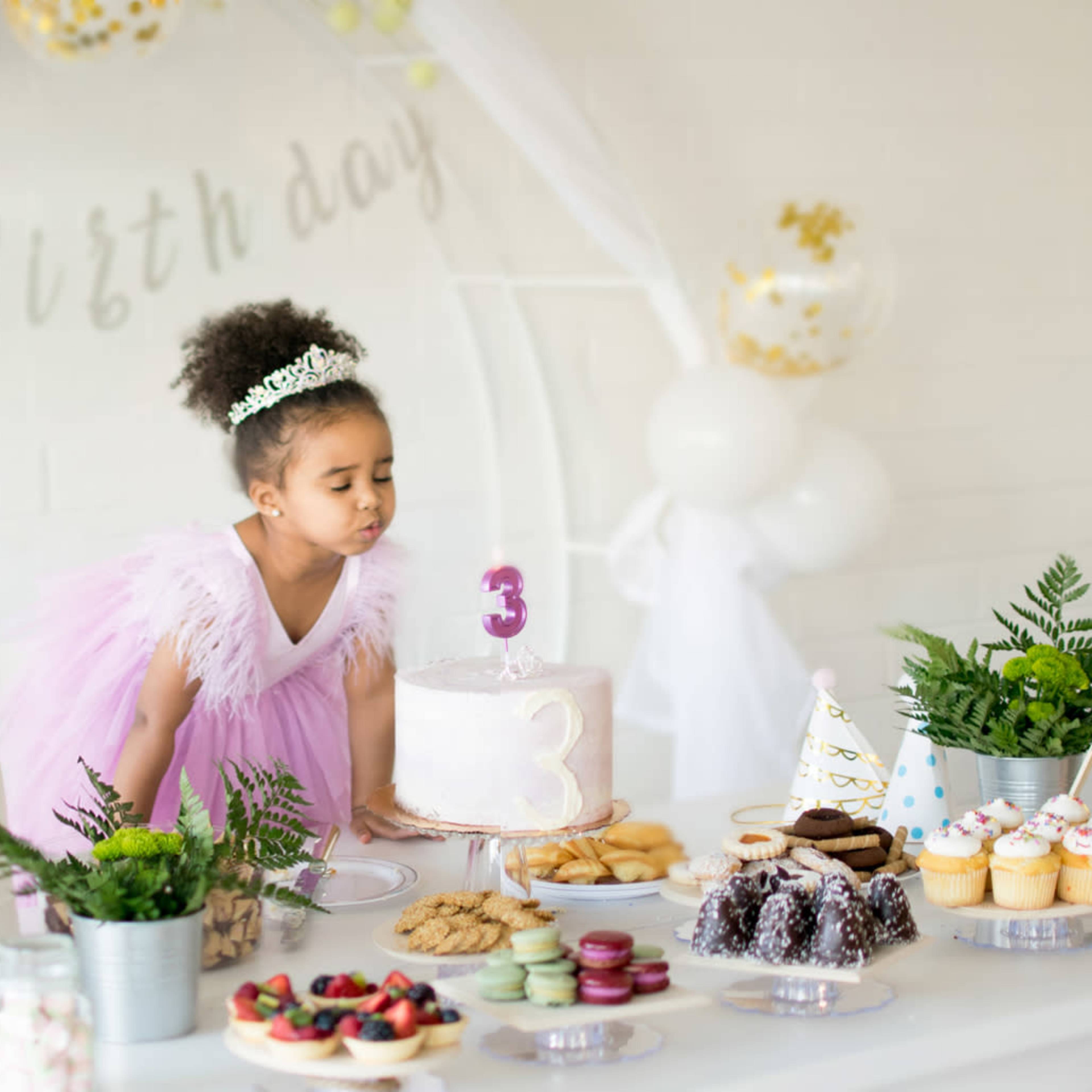 A girl in a pink dress leans over a birthday cake on a decorated table filled with various desserts and party decorations.