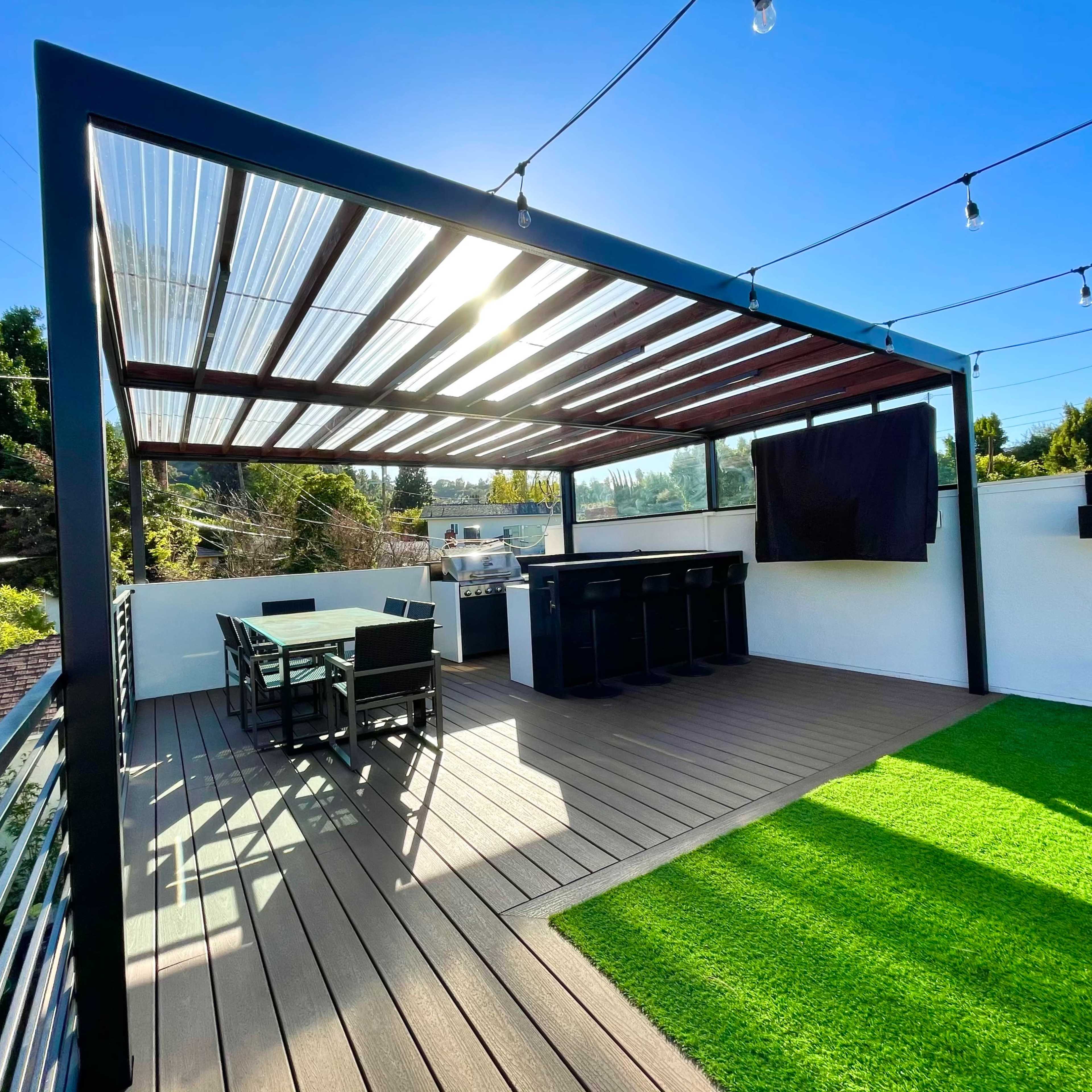 A modern outdoor patio features a wooden deck with a dining table and bar area, surrounded by greenery and string lights overhead.