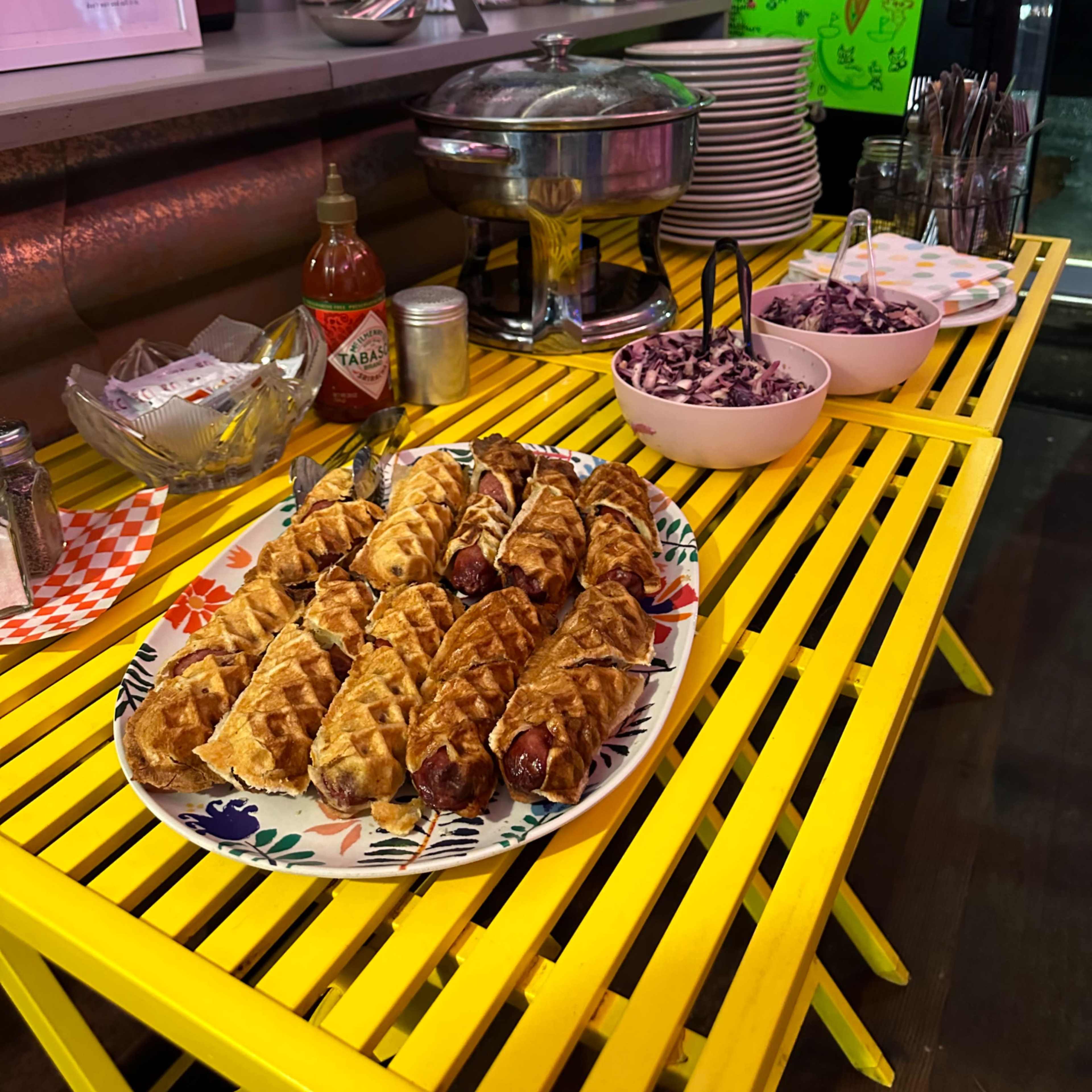 A colorful display of food, including wrapped snacks and bowls of coleslaw, is set on a yellow table in a vibrant kitchen.