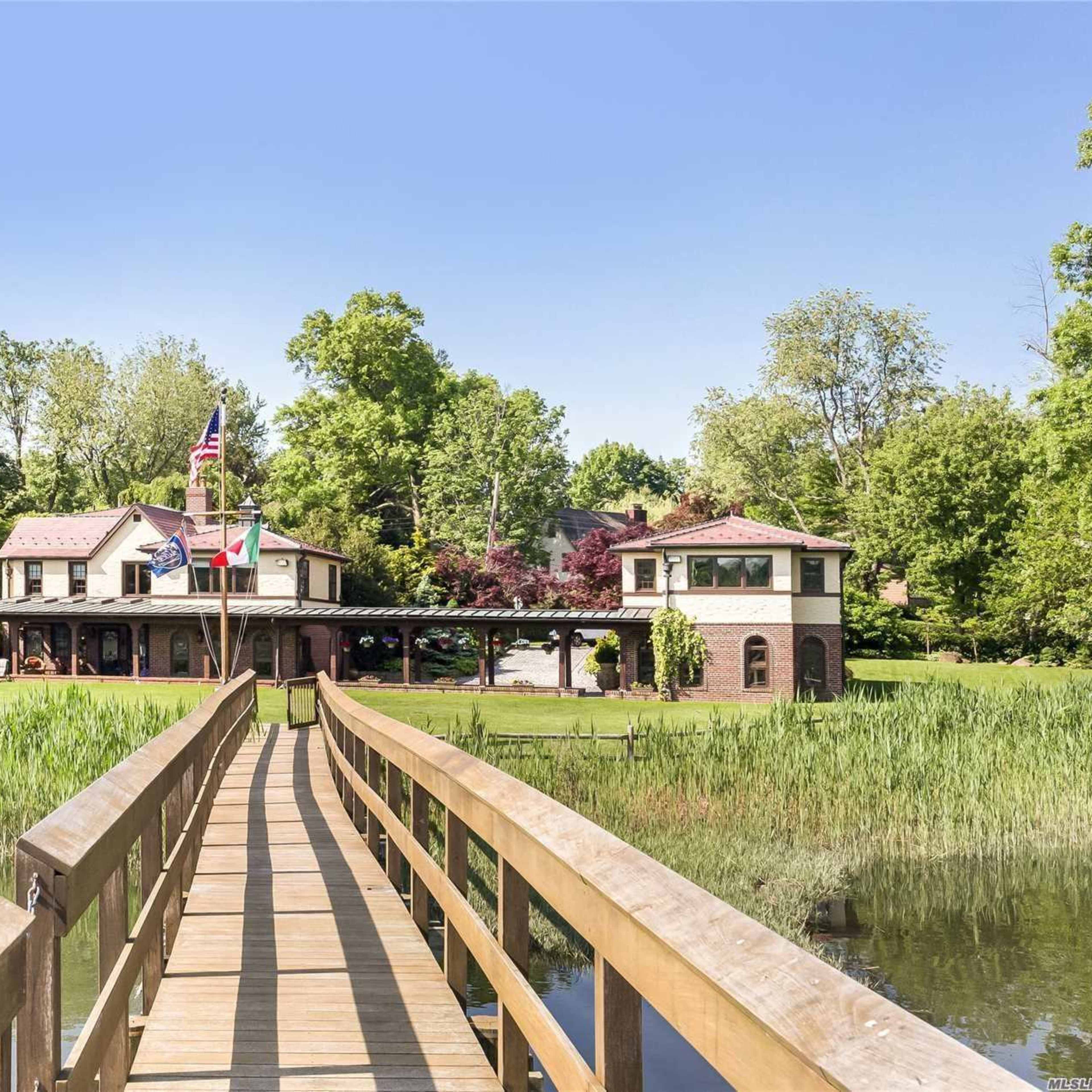 A wooden walkway leads to a large house surrounded by trees and greenery by a calm body of water.