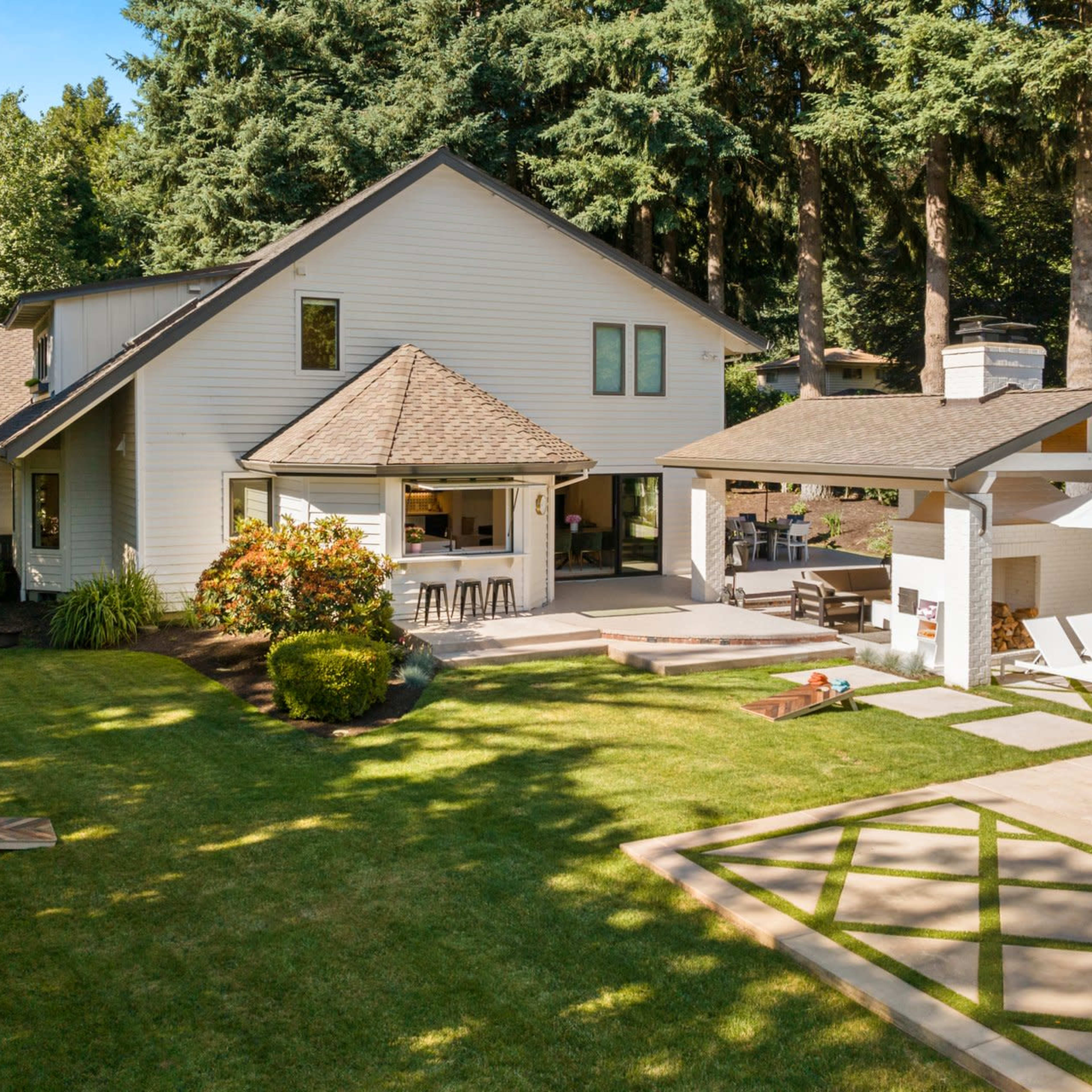 A two-story white house with a patio and outdoor seating area, surrounded by lush greenery and a swimming pool.