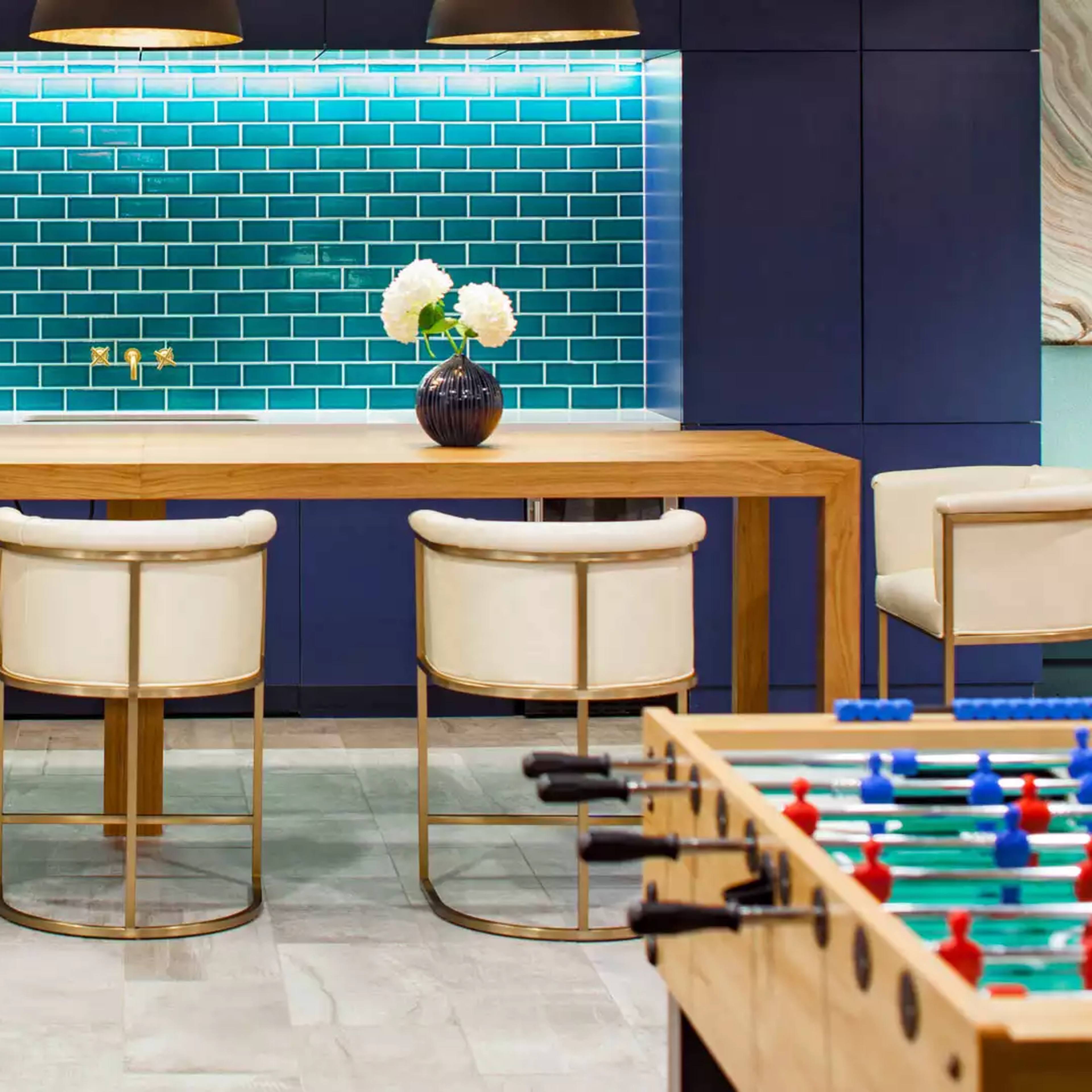 A modern lounge area features a wooden table with three white stools, blue tile backsplash, and a foosball table on either side.