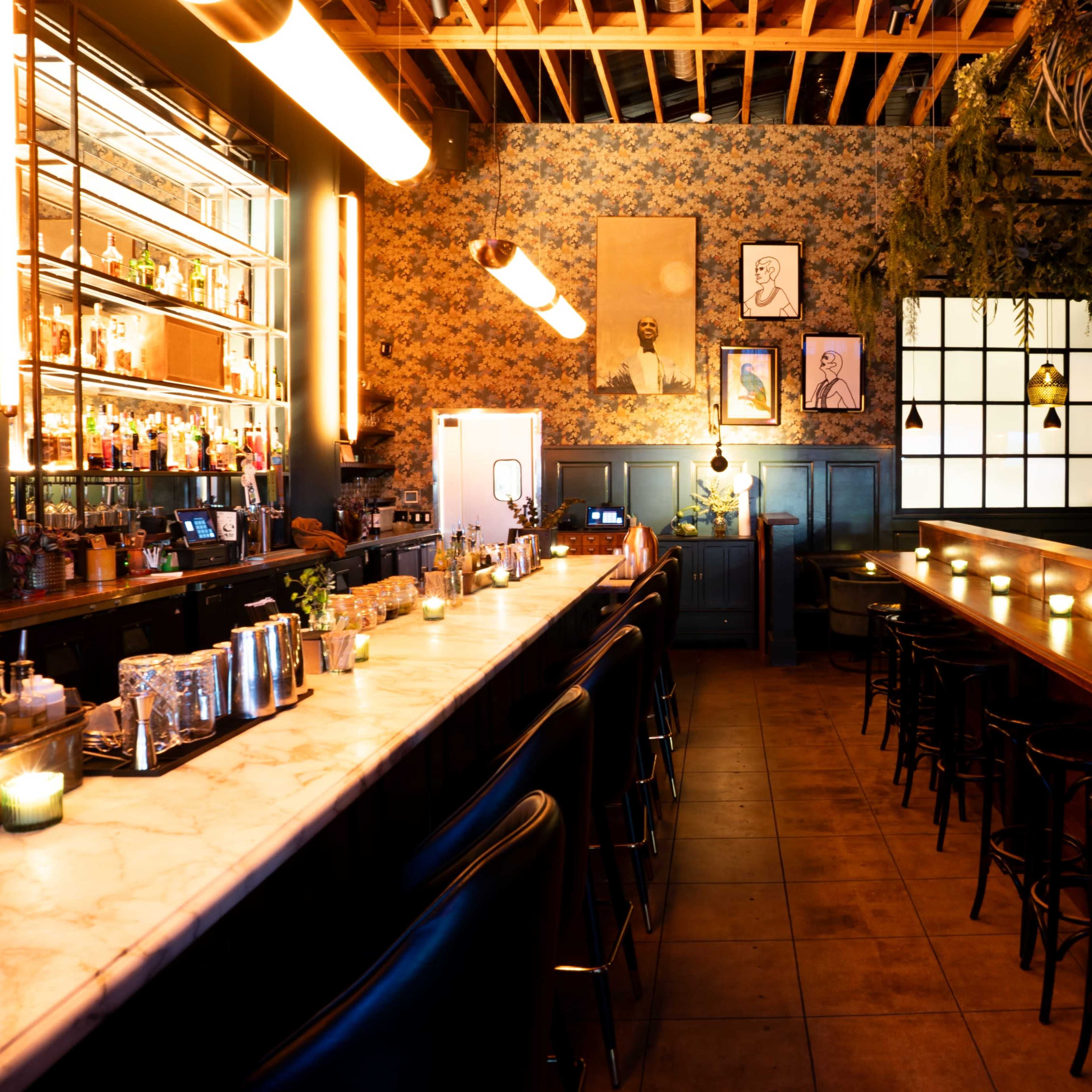 An elegantly designed bar interior with a long marble countertop, high stools, and ambient lighting, complemented by floral wallpaper and decorative elements on the walls.
