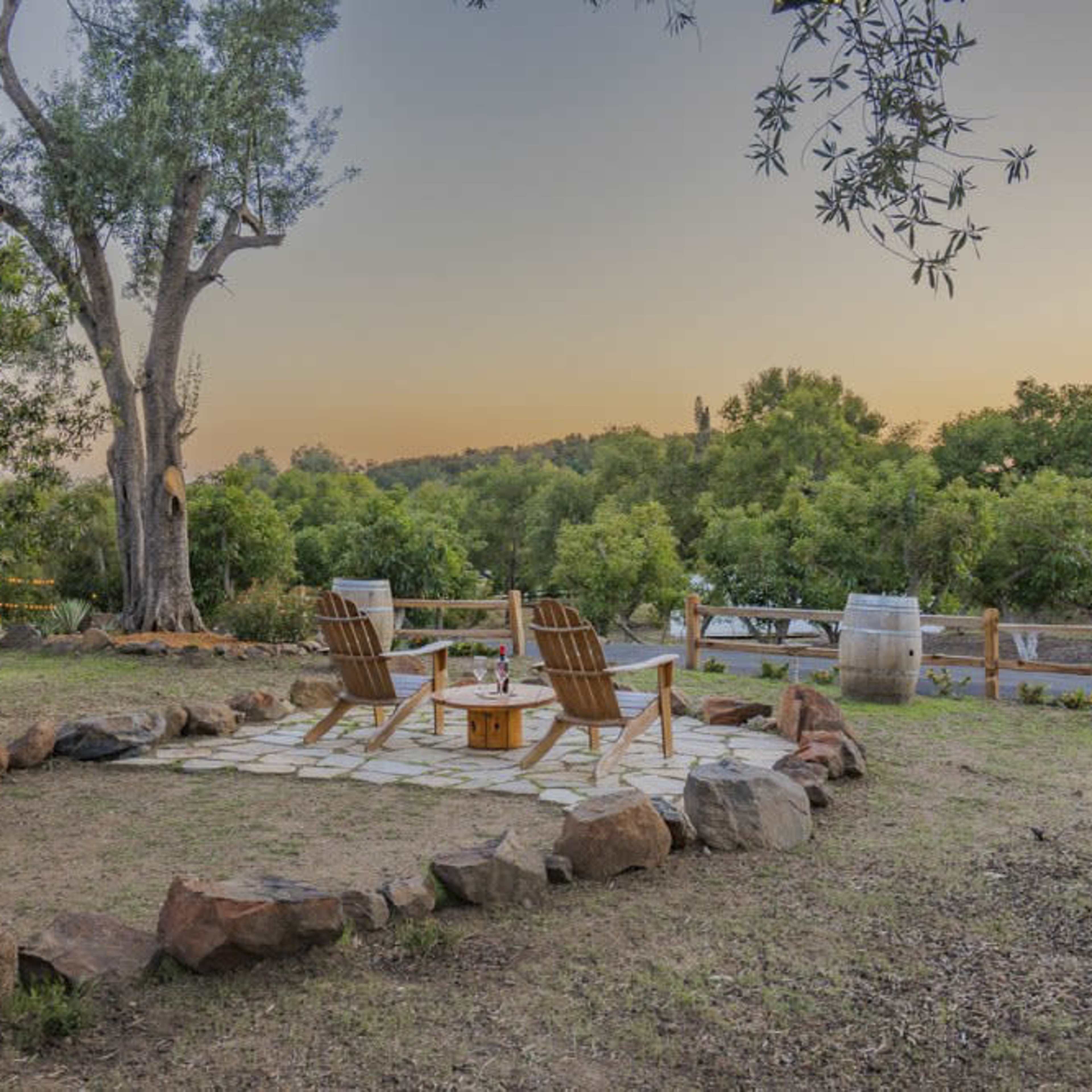 A stone patio with two wooden chairs surrounds a fire pit in a garden area, bordered by trees and greenery.
