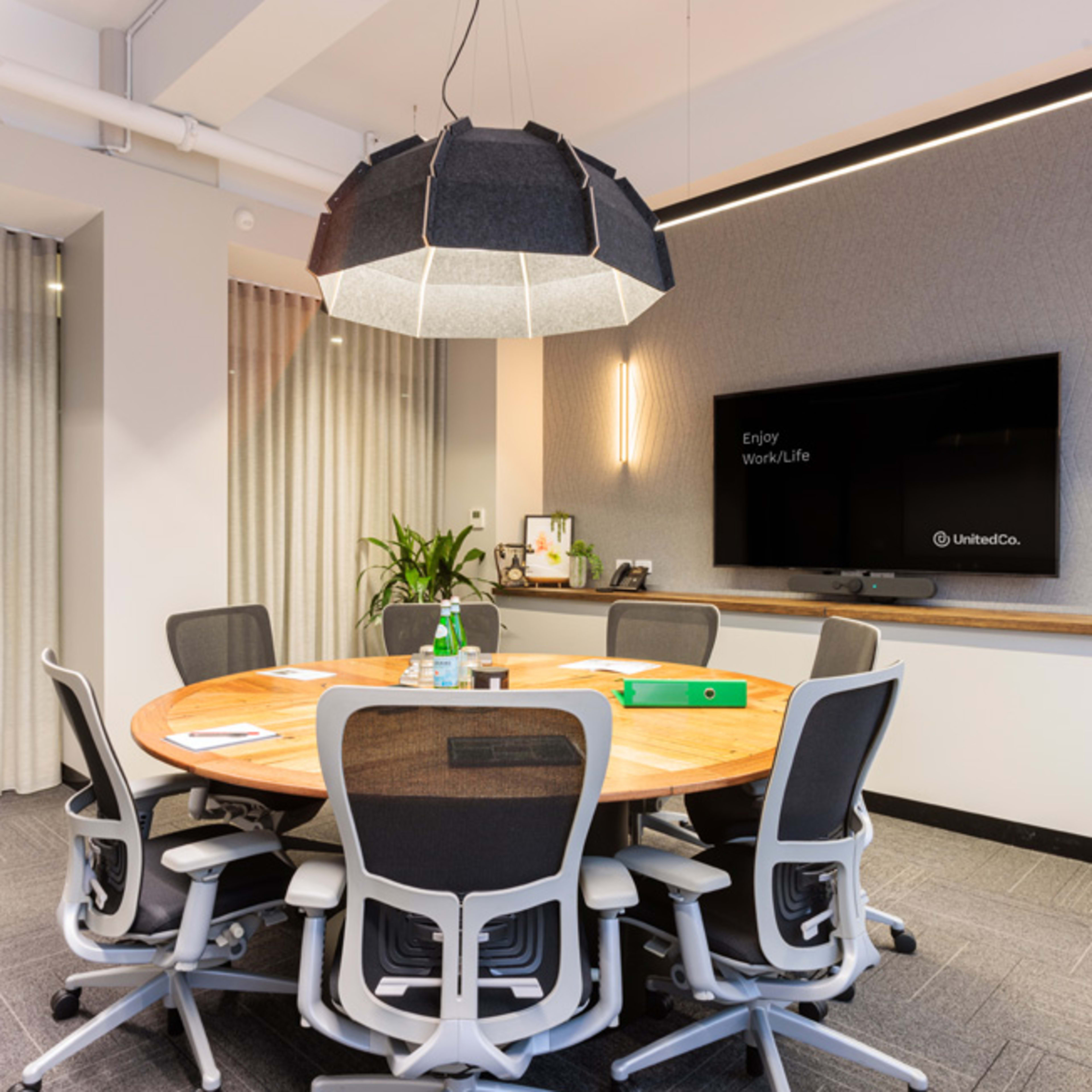 A modern conference room with a circular wooden table surrounded by ergonomic chairs, a large pendant light overhead, and a TV mounted on the wall.