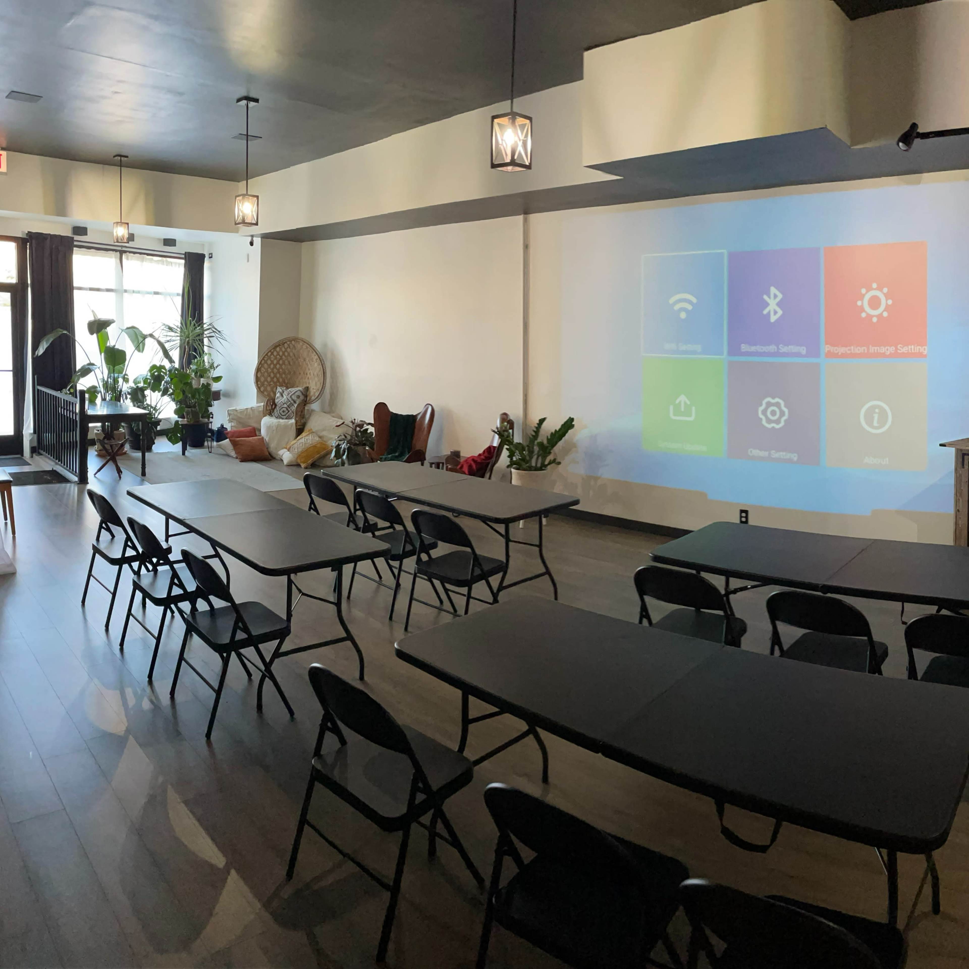A room with several tables arranged in a U-shape, featuring a large projection screen on one wall and plants decoratively placed around the space.