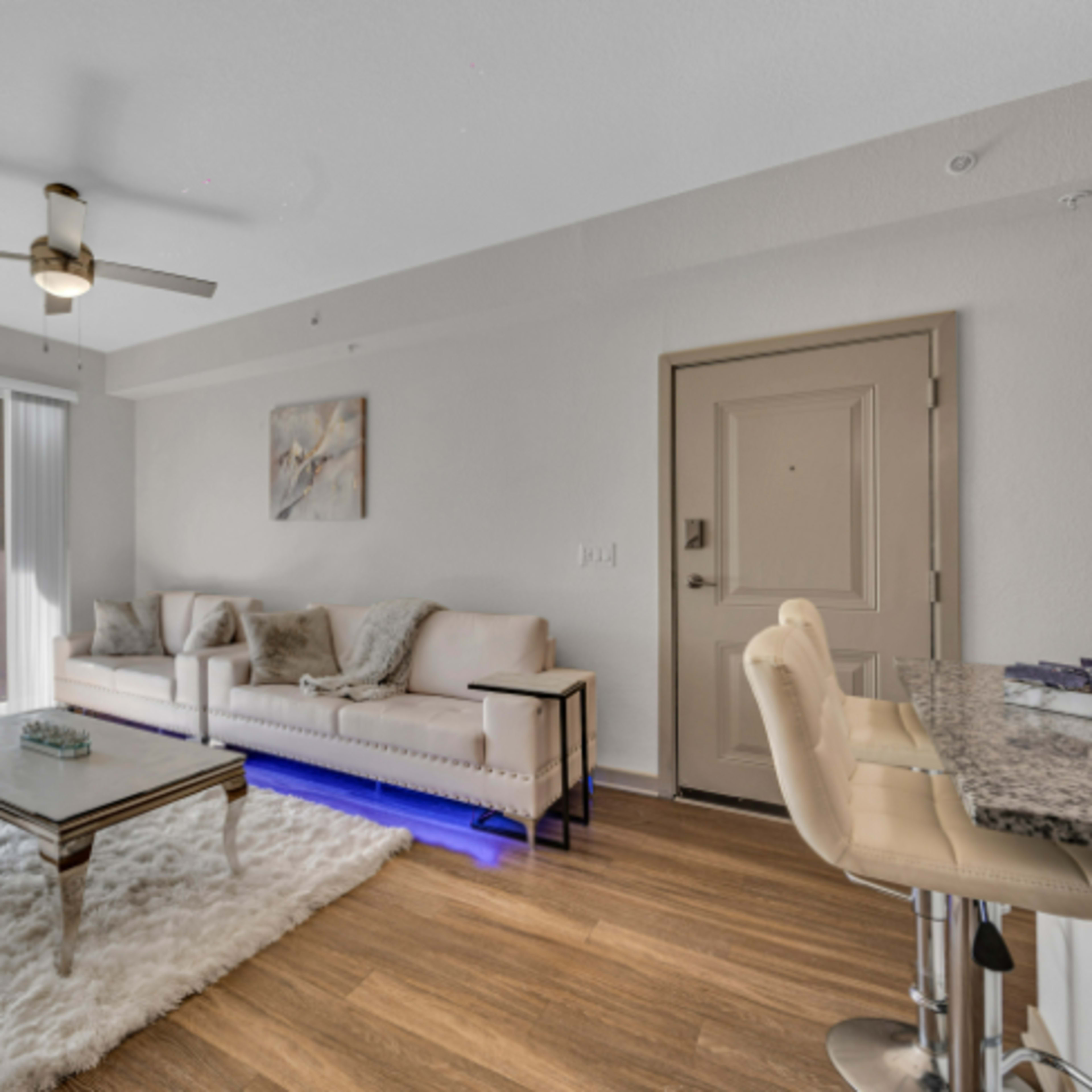 A modern living room features a light-colored sofa, a glass coffee table on a rug, and a kitchen bar with stools, illuminated by natural light from large windows.