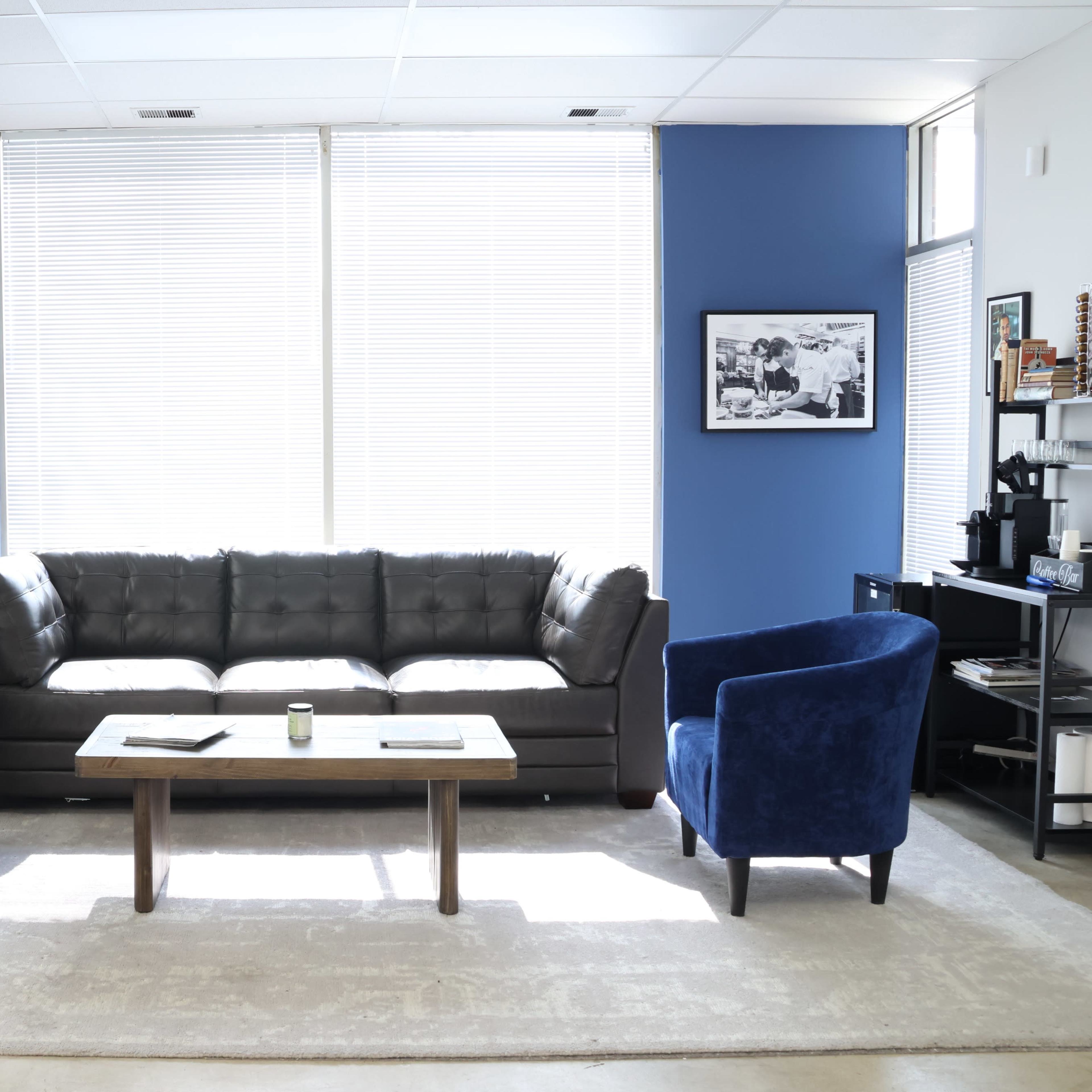 A modern lounge area with a brown leather sofa, two blue armchairs, a wooden coffee table, and large windows with white blinds.
