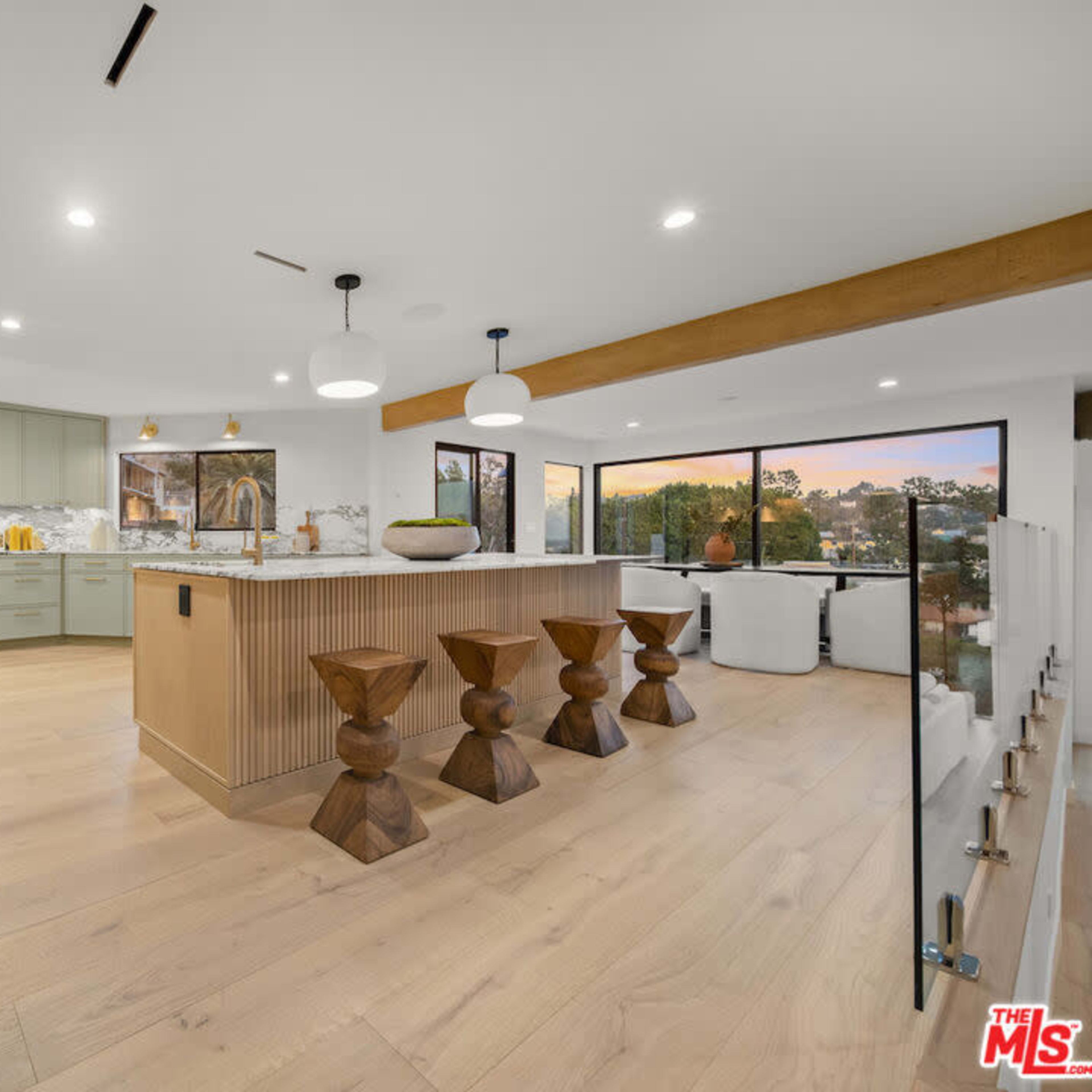 A modern kitchen with light wood flooring, a large central island, and large windows that provide a view of the outdoors.