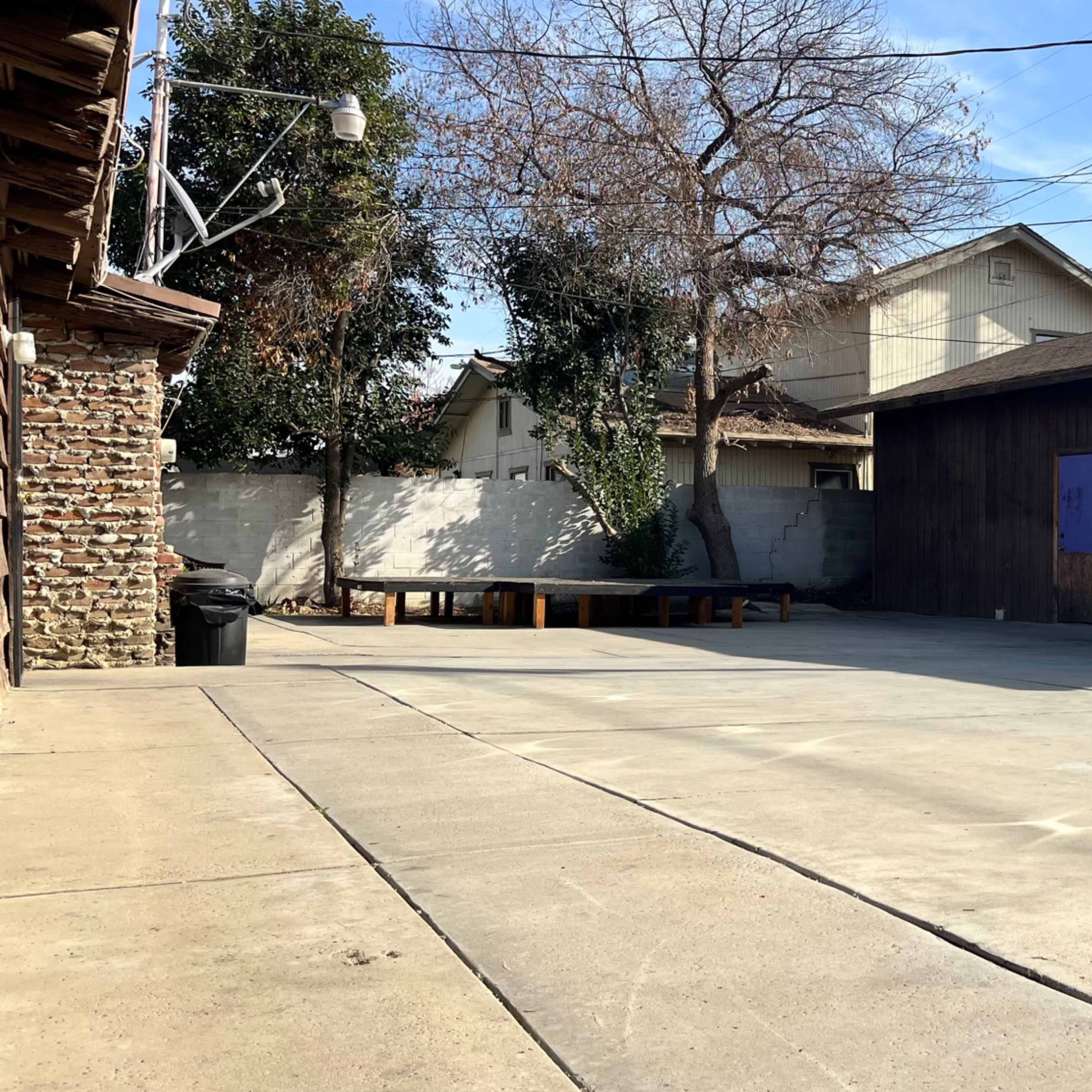 An outdoor area featuring a concrete surface with a wooden building on one side and a table with benches on the opposite side, surrounded by trees and utility poles.