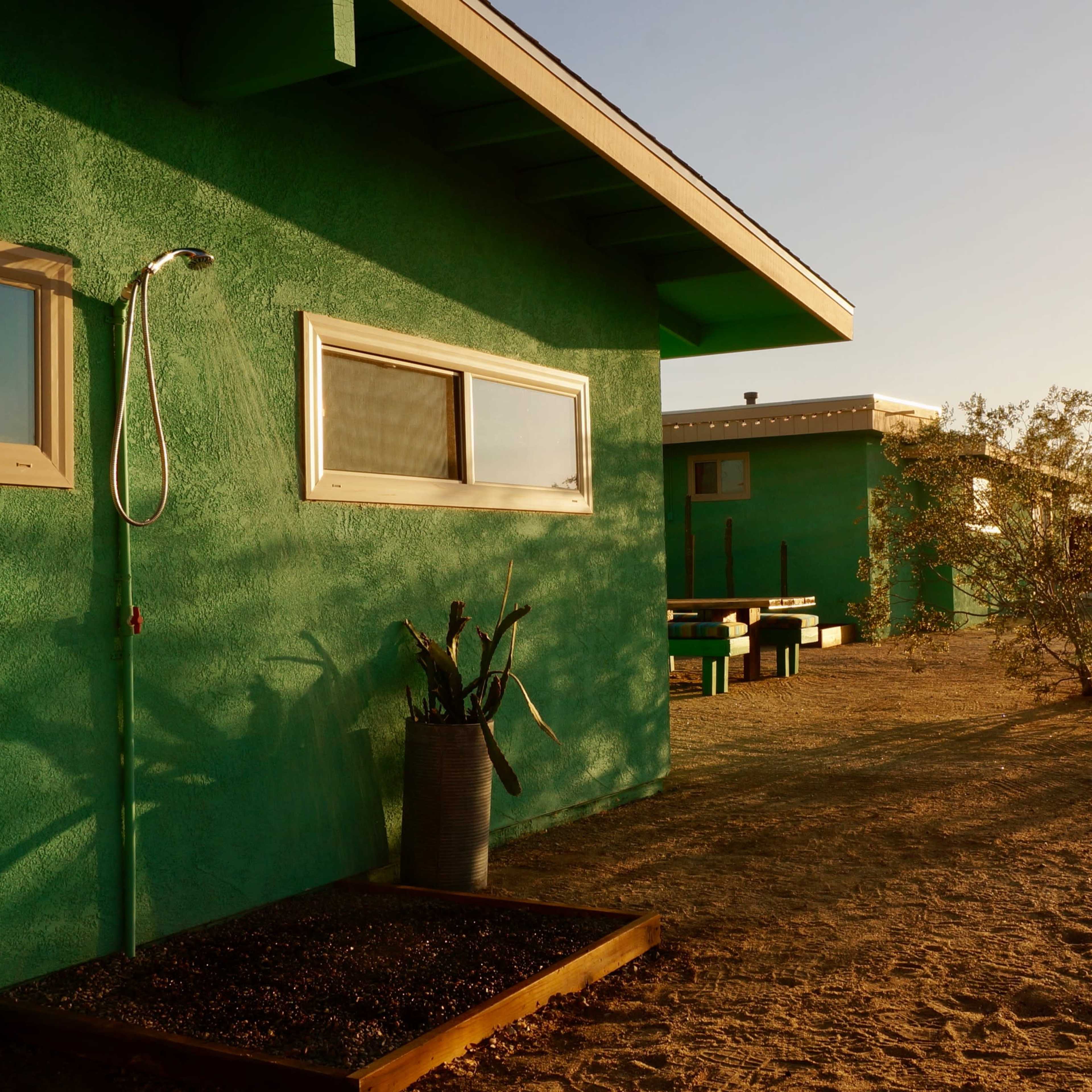 A green house with a hose attached to its side is set against a desert landscape with sparse vegetation and a clear sky.