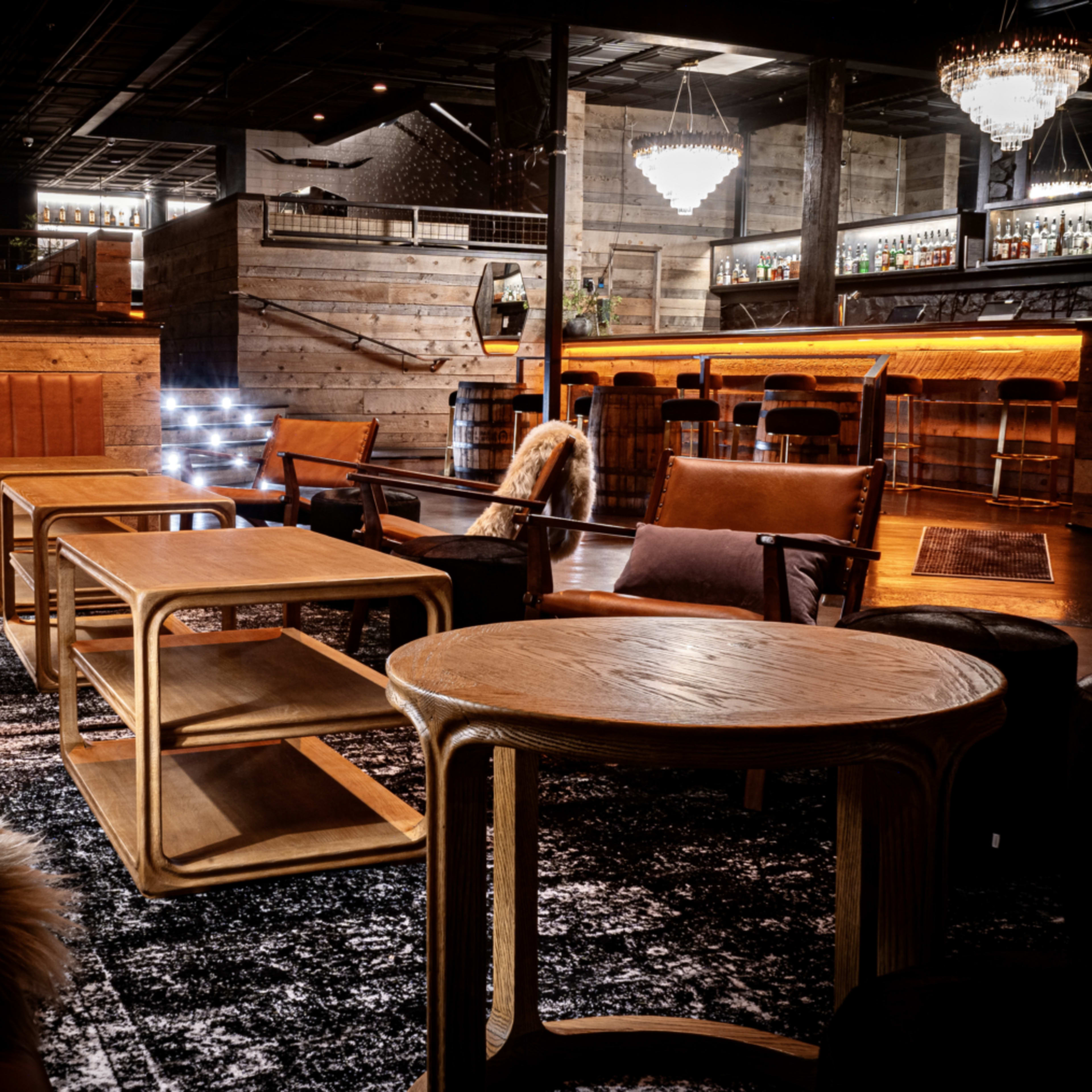 A stylish lounge area with wooden furniture, a patterned rug, and a well-lit bar in the background.