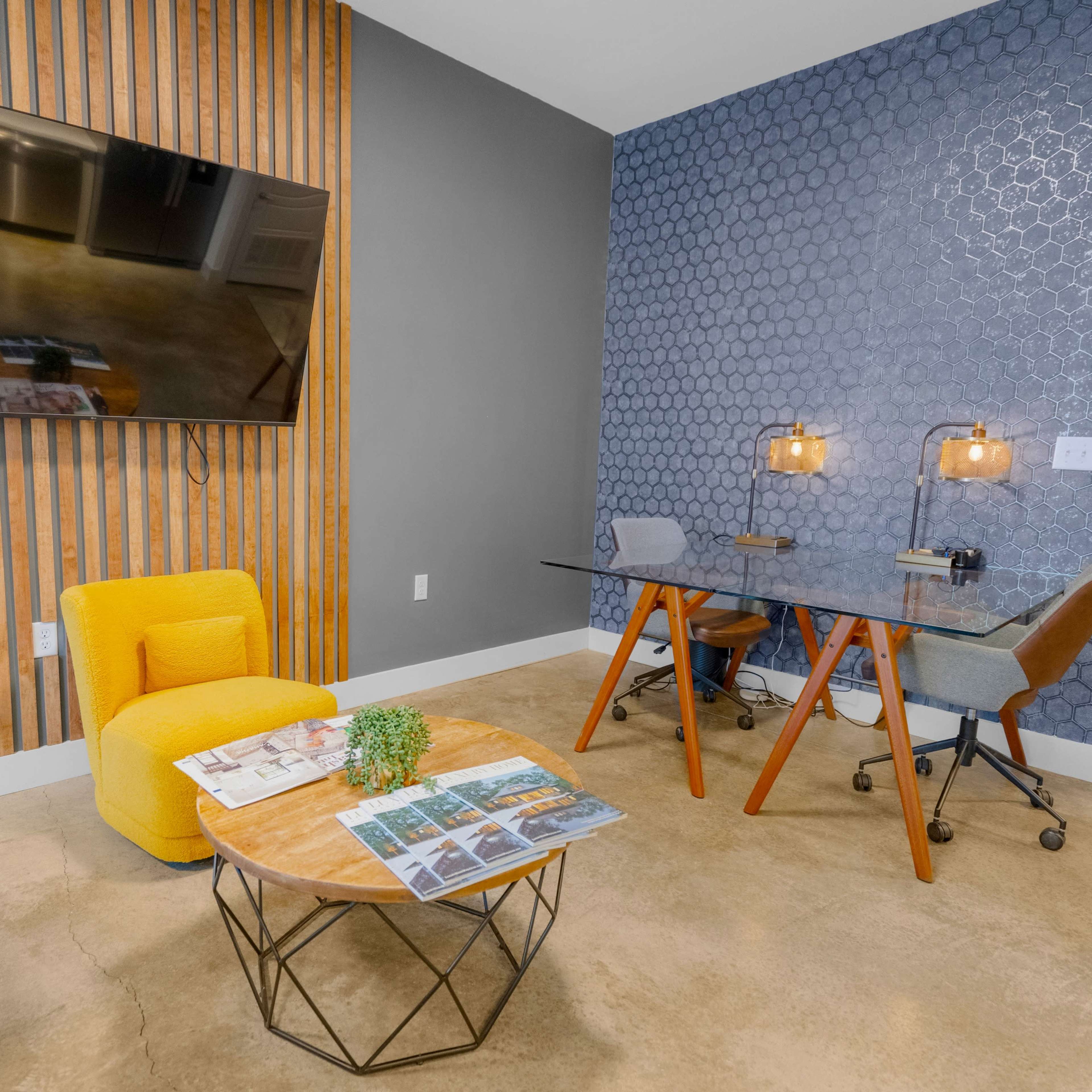 A modern office space featuring a yellow armchair, a coffee table with magazines, a glass desk with task lamps, and a television mounted on a wooden wall.