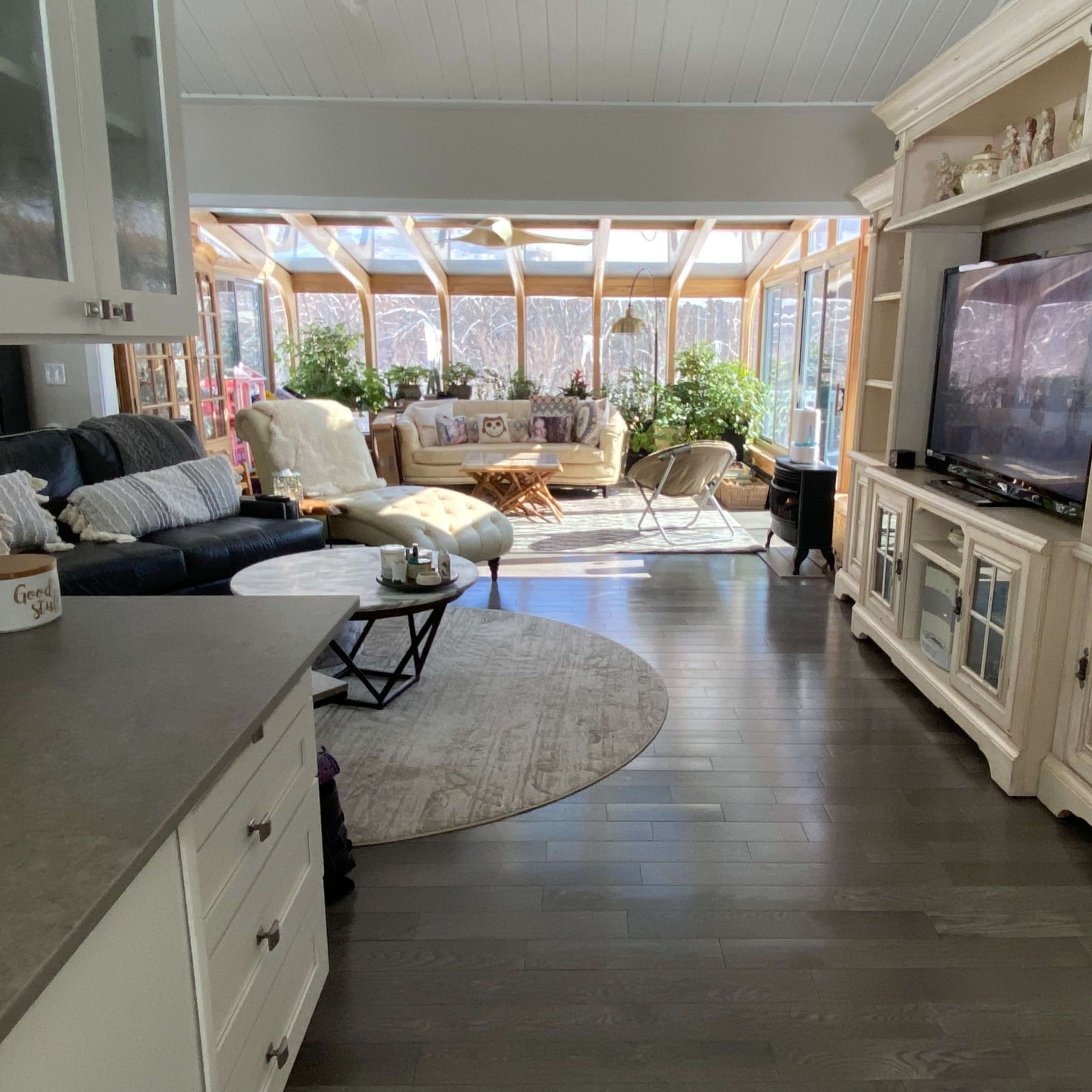 A spacious living area with light-colored furniture, a large television, and a sunroom filled with plants and natural light.