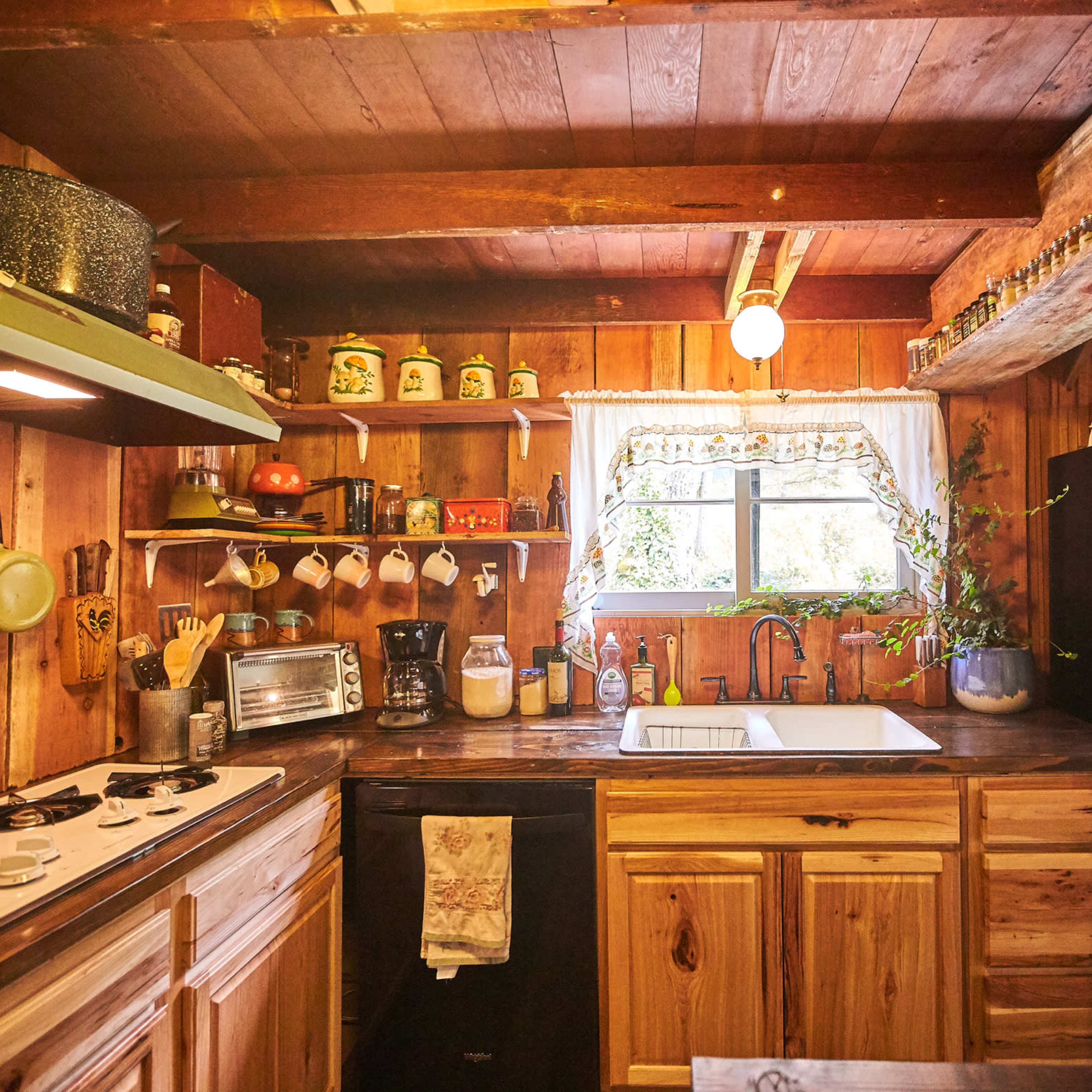 A rustic kitchen with wooden walls and cabinetry, featuring a sink beneath a window, a gas stove, and various kitchen utensils on display.