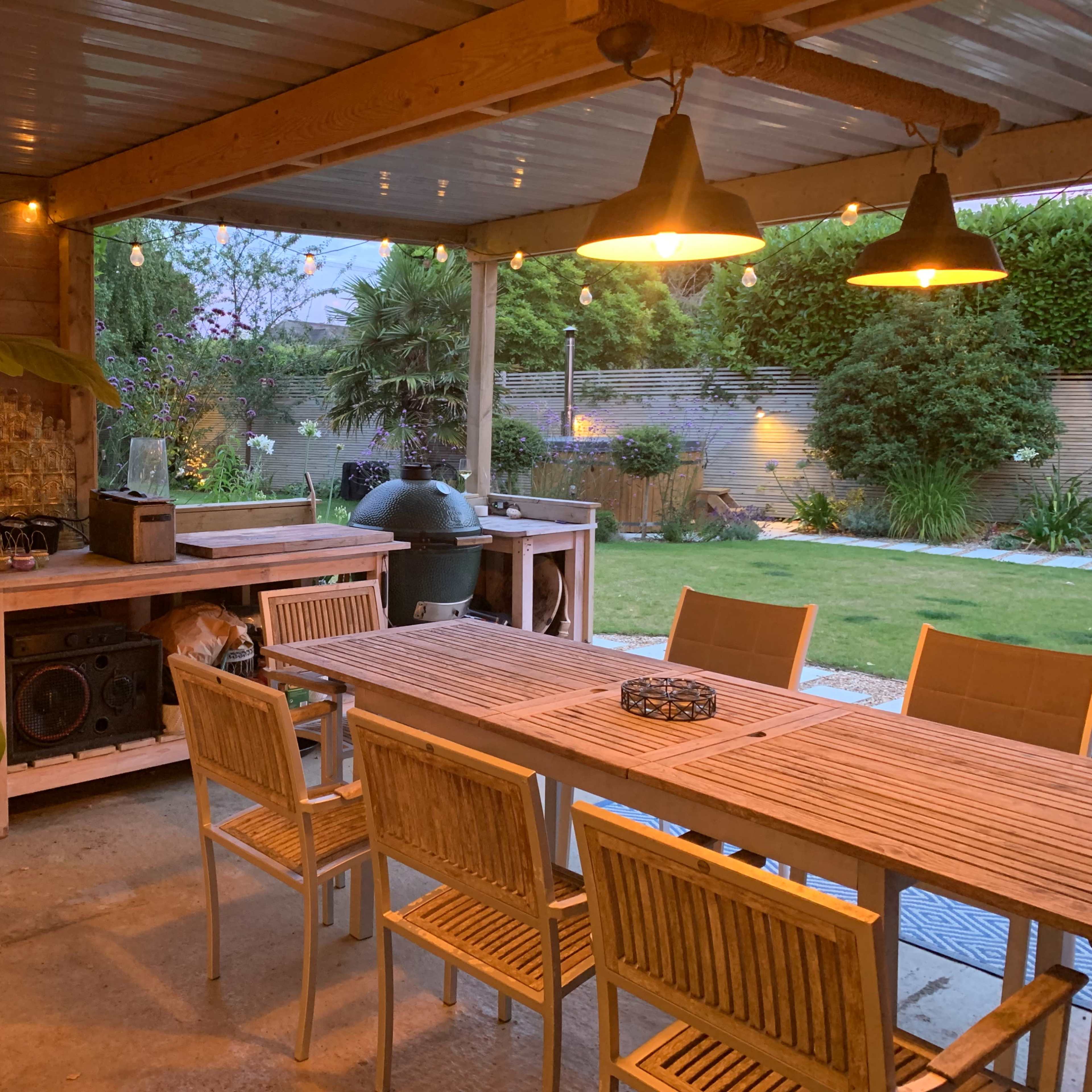 A well-designed outdoor patio features a wooden dining table surrounded by chairs, a grill area, and a landscaped garden in the background.