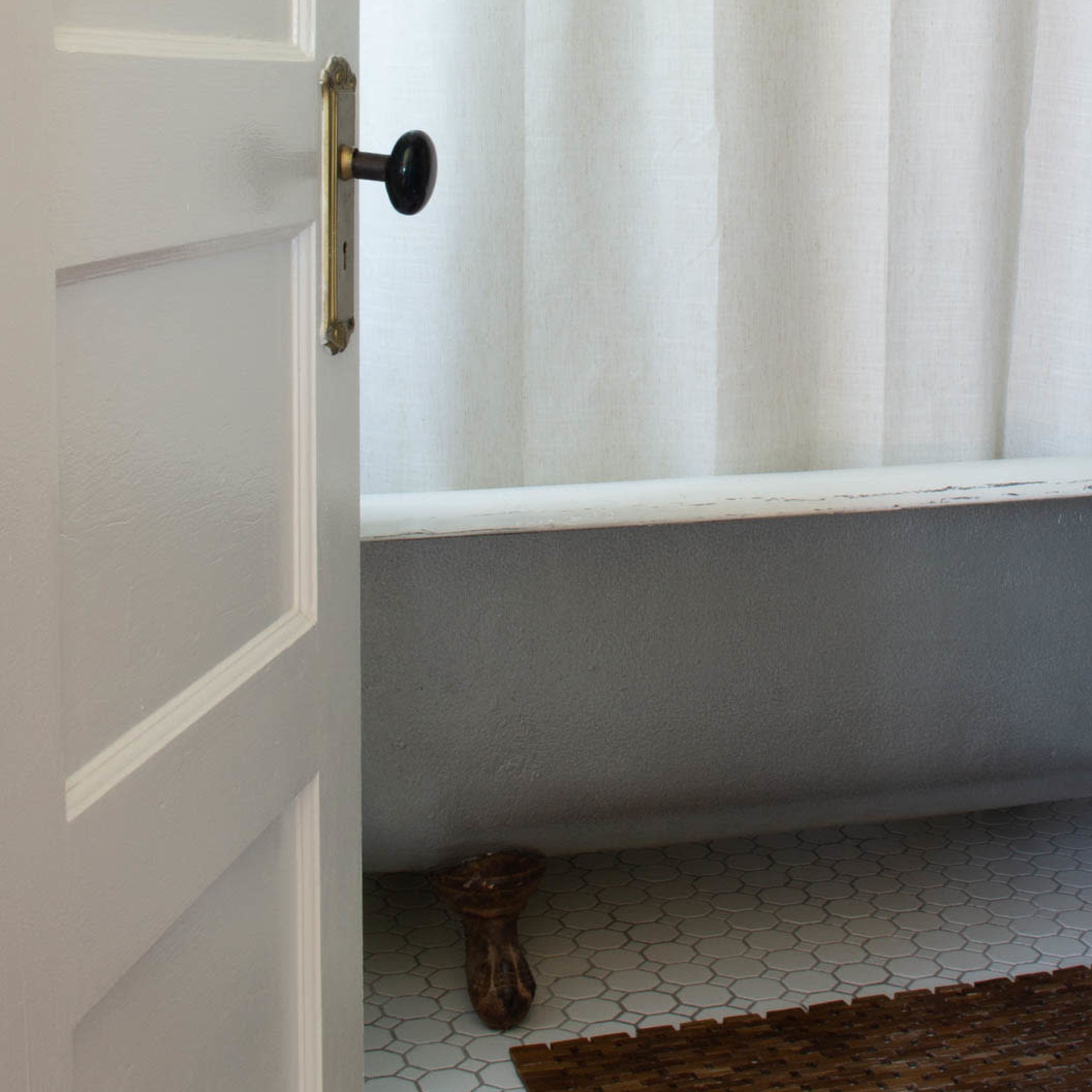 A white door partially opens to reveal a bathroom with a patterned shower curtain, a bathtub, and a tiled floor featuring a brown mat.