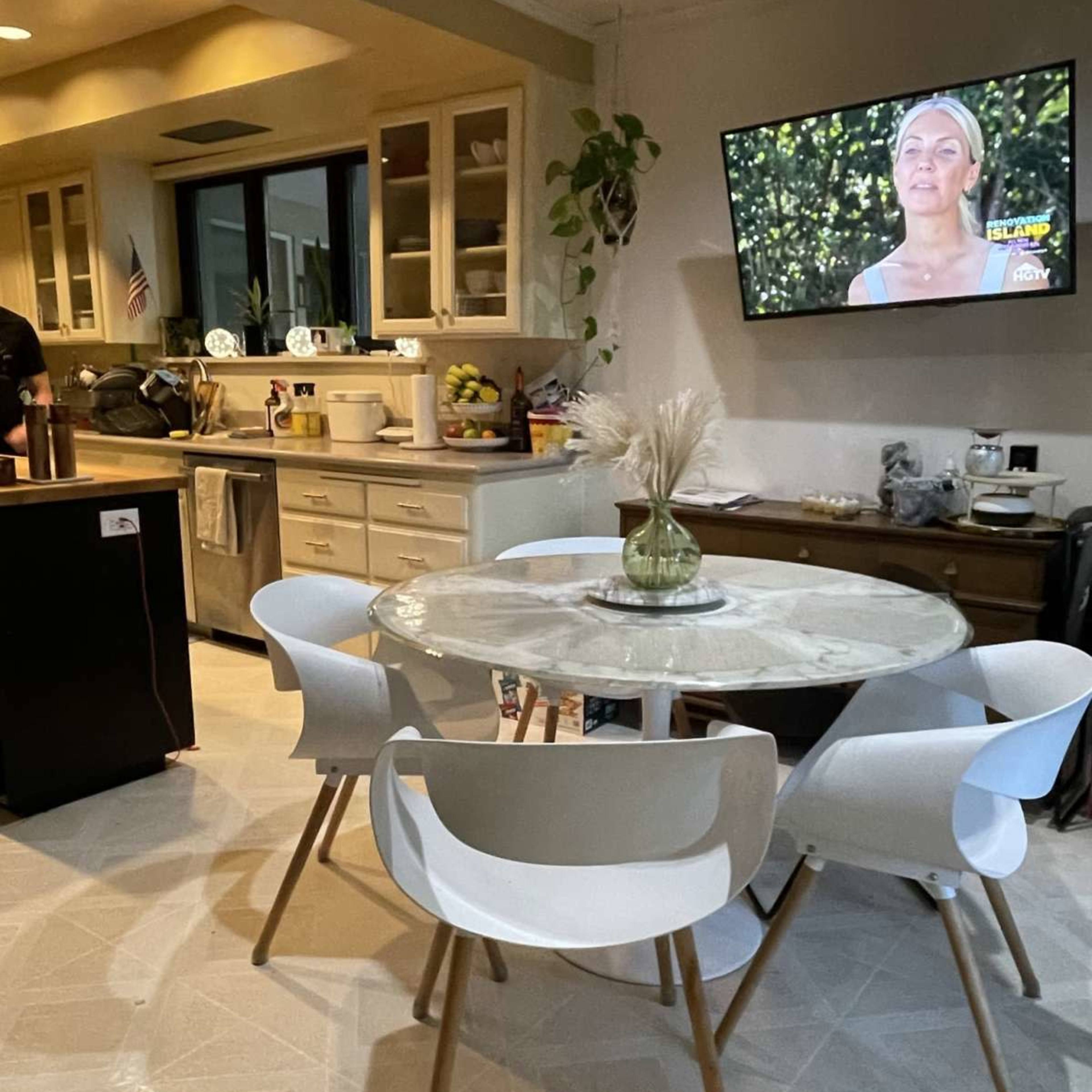 A modern kitchen with a round dining table and white chairs, featuring a television screen displaying a woman in a well-lit room.