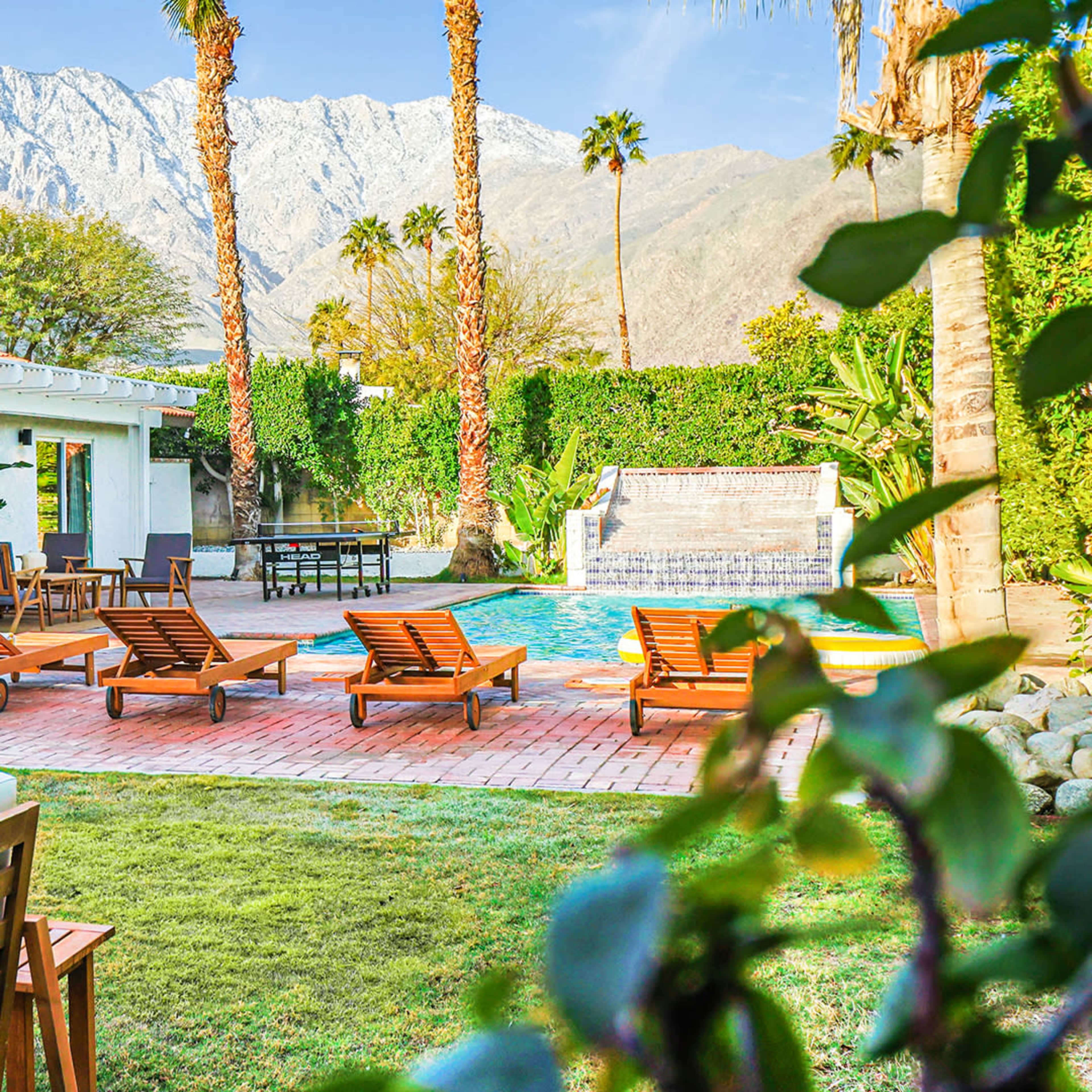 A modern backyard features a swimming pool with lounge chairs, surrounded by palm trees and mountains in the background.