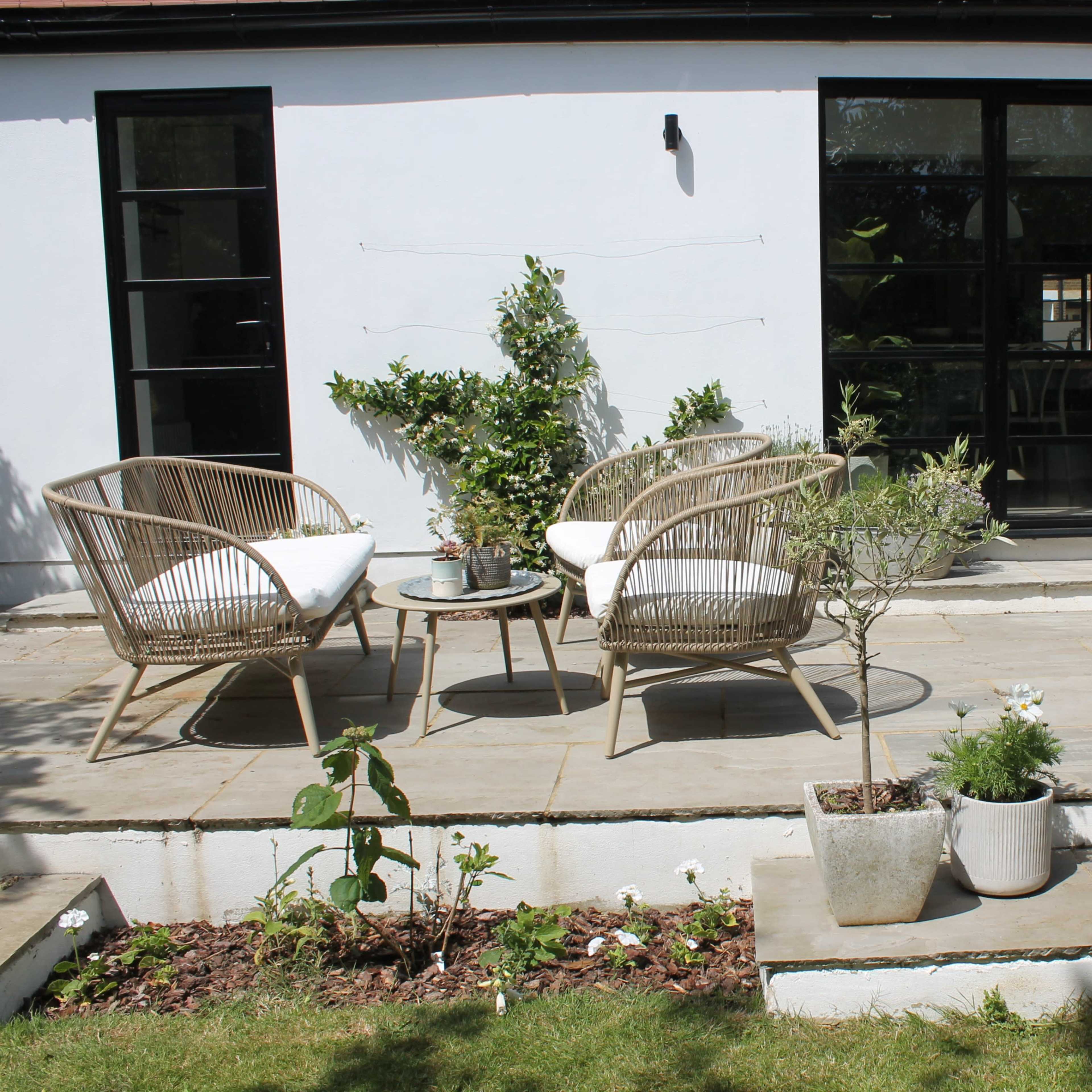 A modern patio with two wicker chairs and a small table, surrounded by planters and a clear view into an interior space through large glass doors.