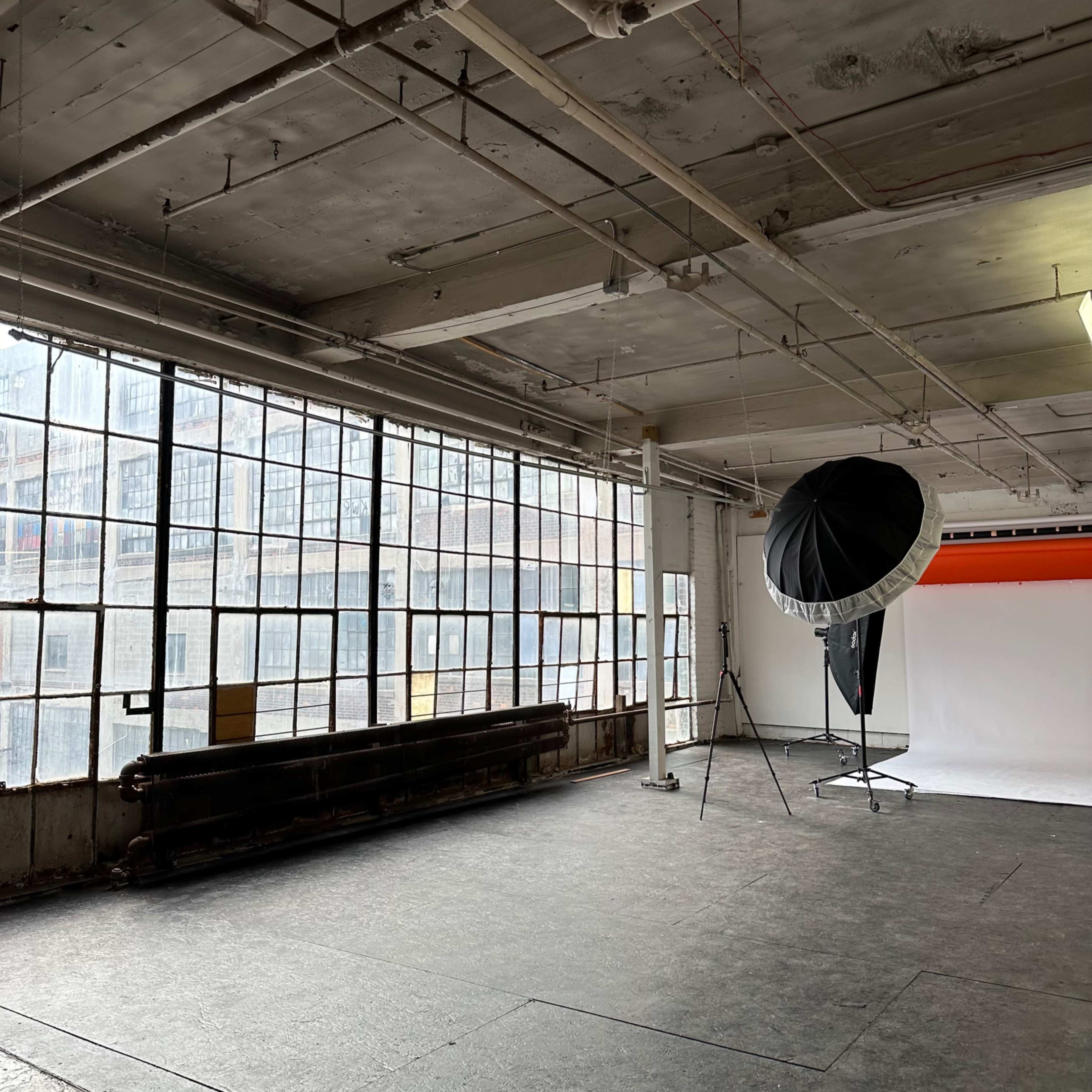 A spacious industrial studio features large windows, a simple backdrop, and a lighting setup with a softbox.