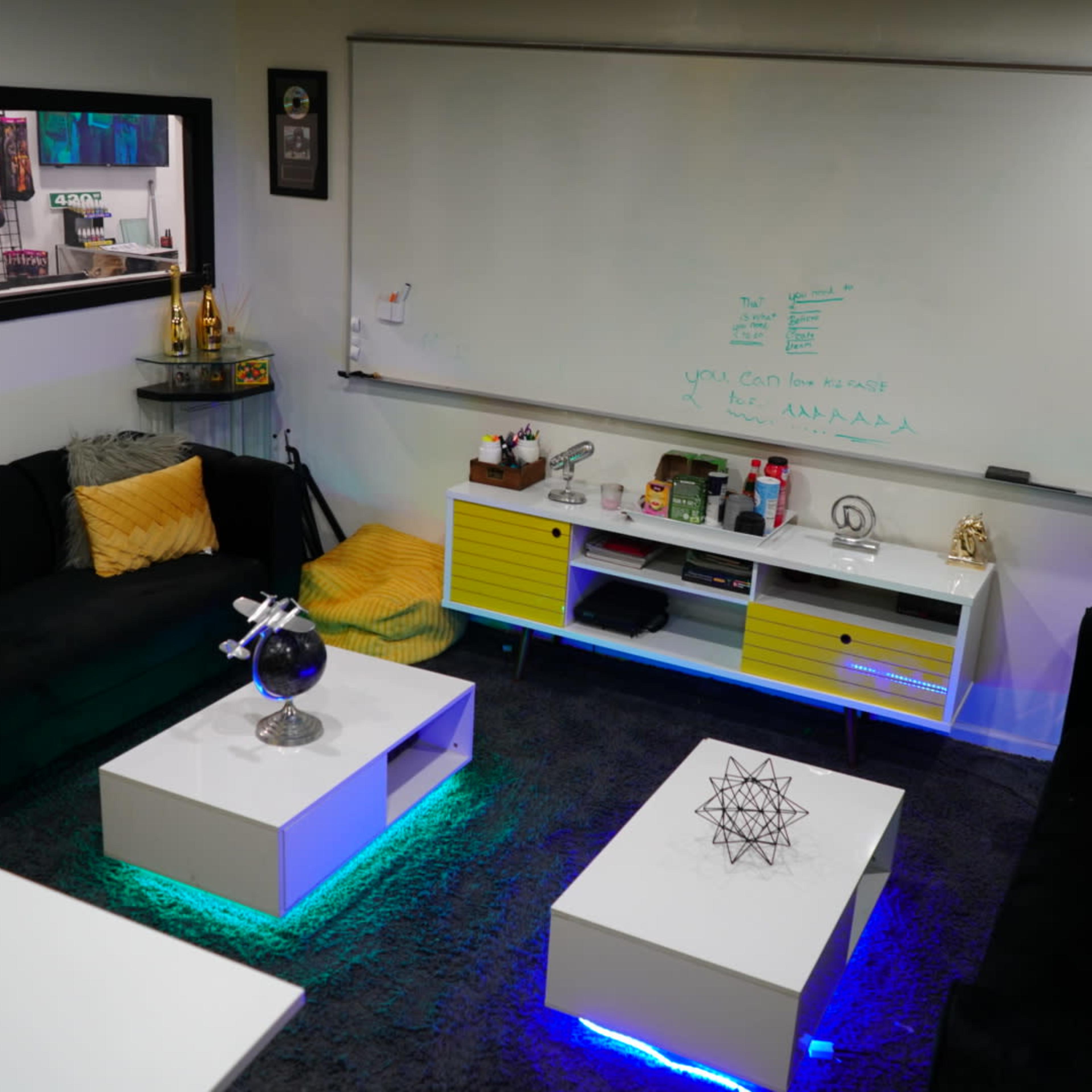 A modern lounge area with black sofas, white coffee tables featuring LED lighting, and a whiteboard mounted on the wall.