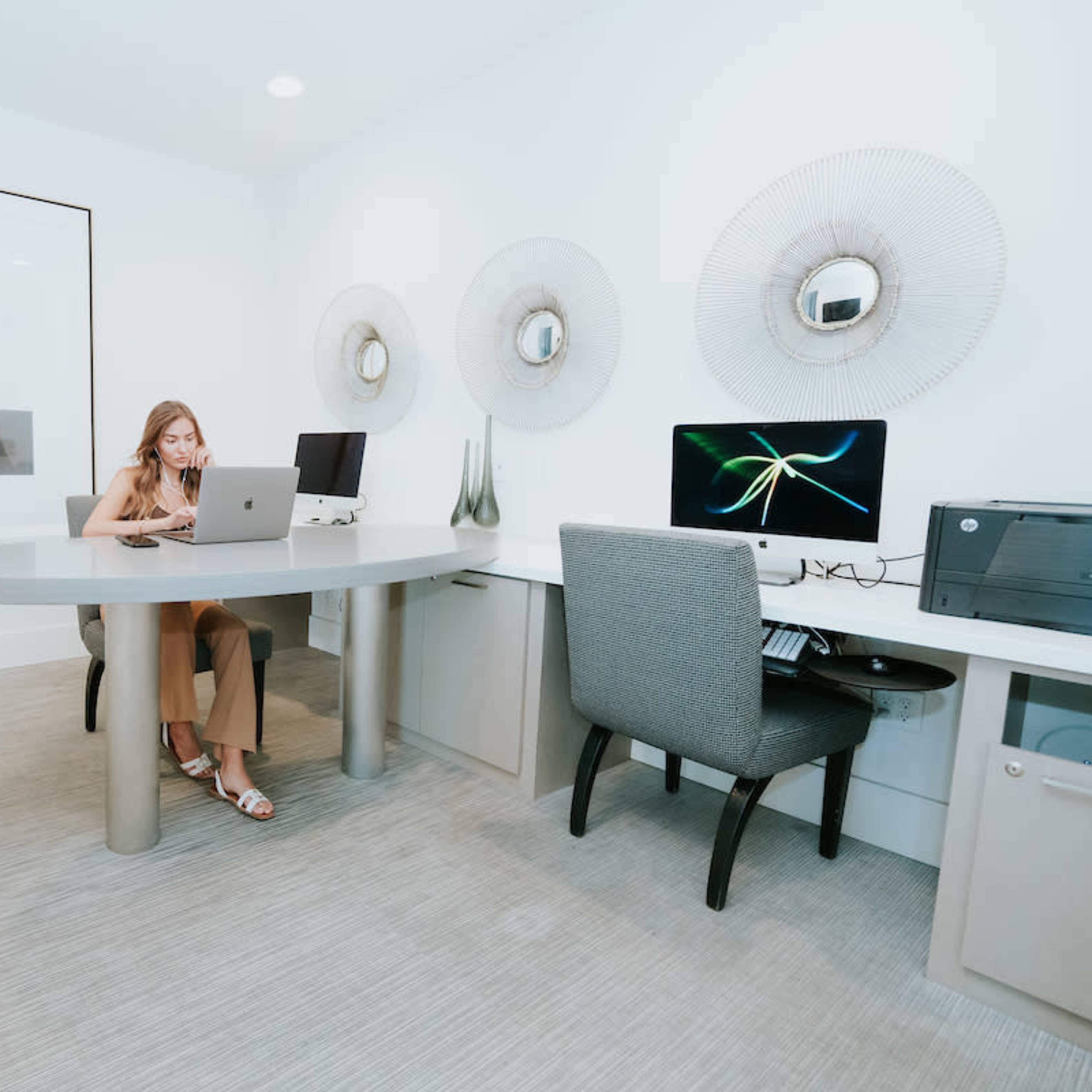 A woman sits at a round desk in a modern office space, working on a laptop, with two computers and decorative wall elements in the background.