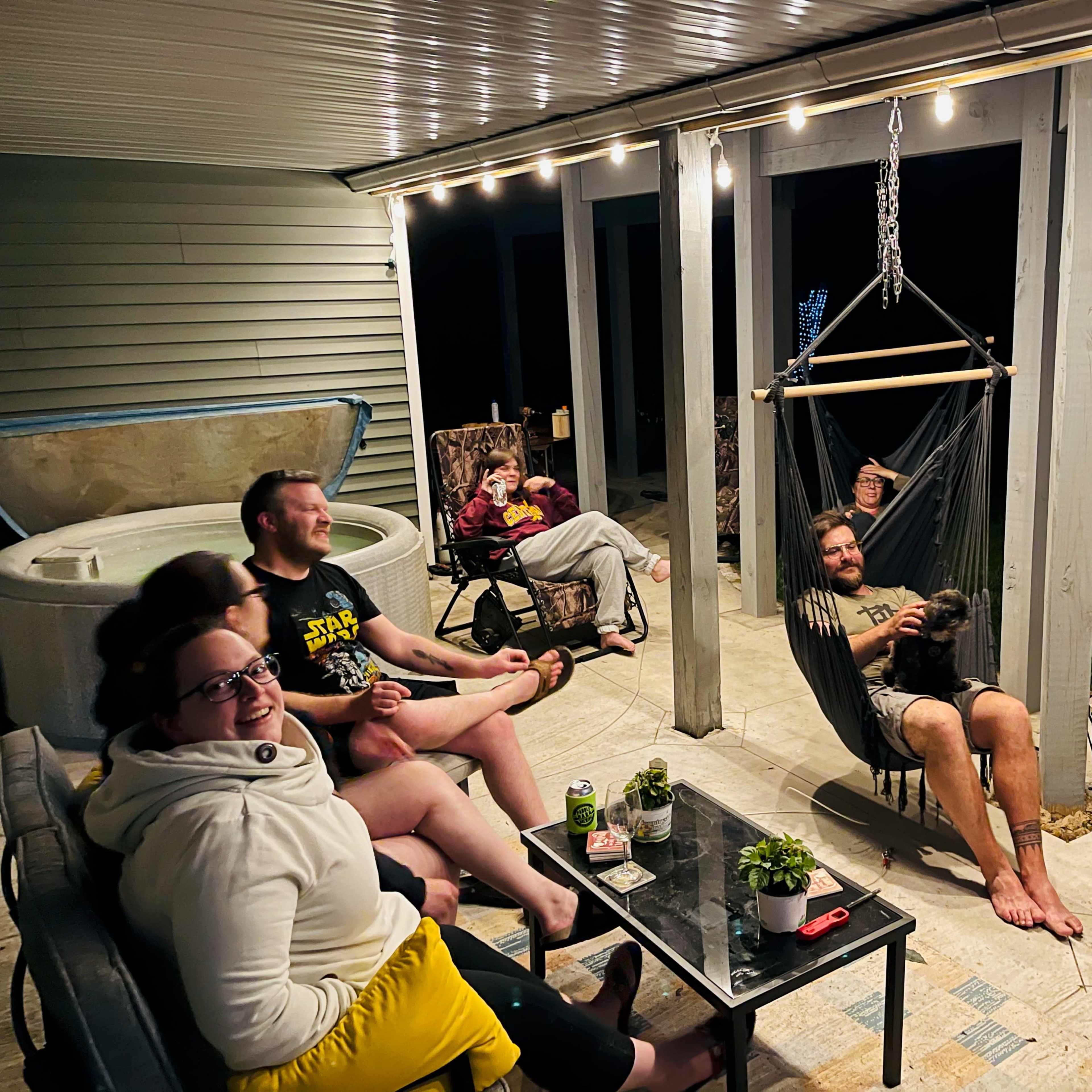 A group of six people sits in a well-lit outdoor space with a hot tub in the background, enjoying each other's company in chairs and a hammock.