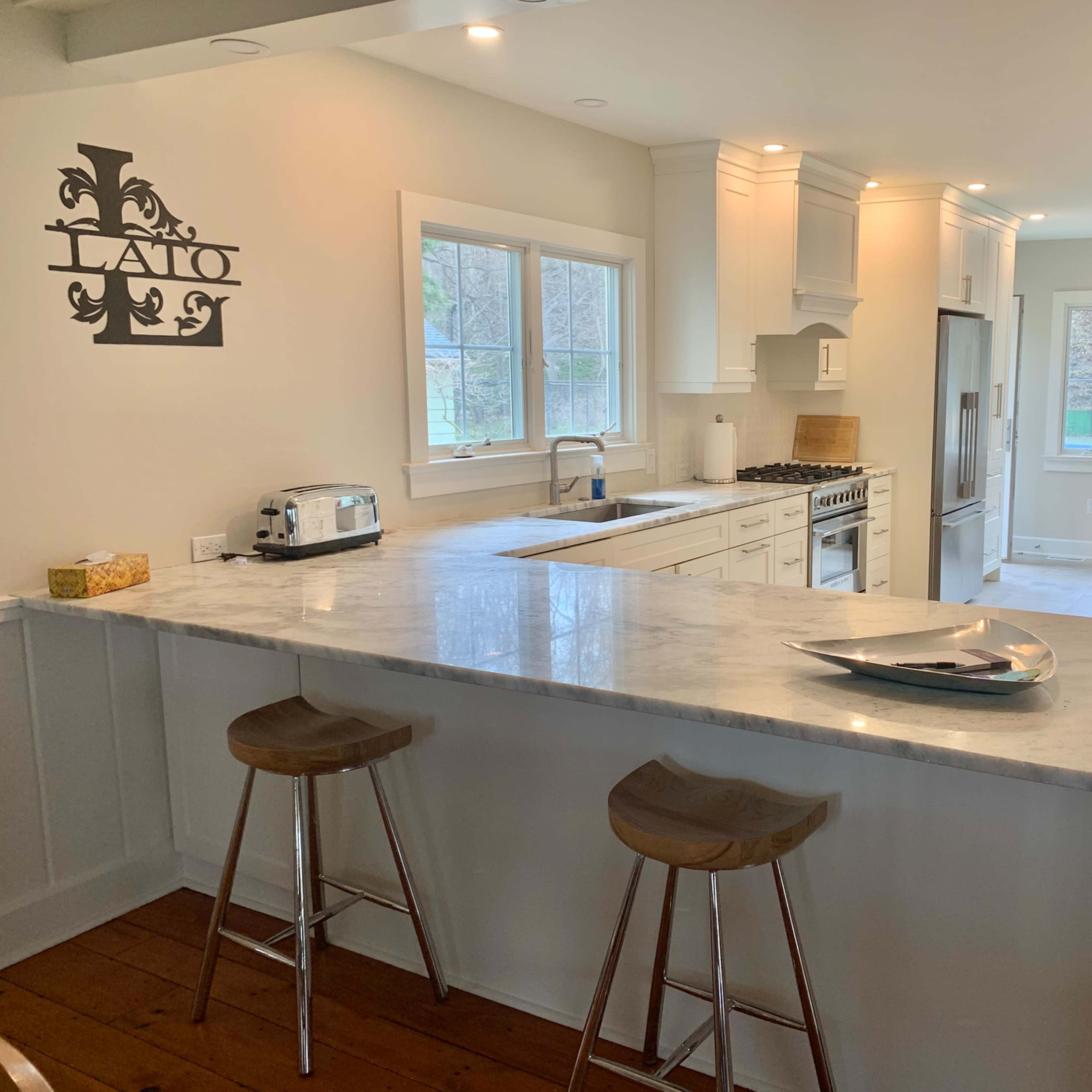 A bright kitchen features white cabinetry, a marble countertop with bar stools, and large windows overlooking a backyard.