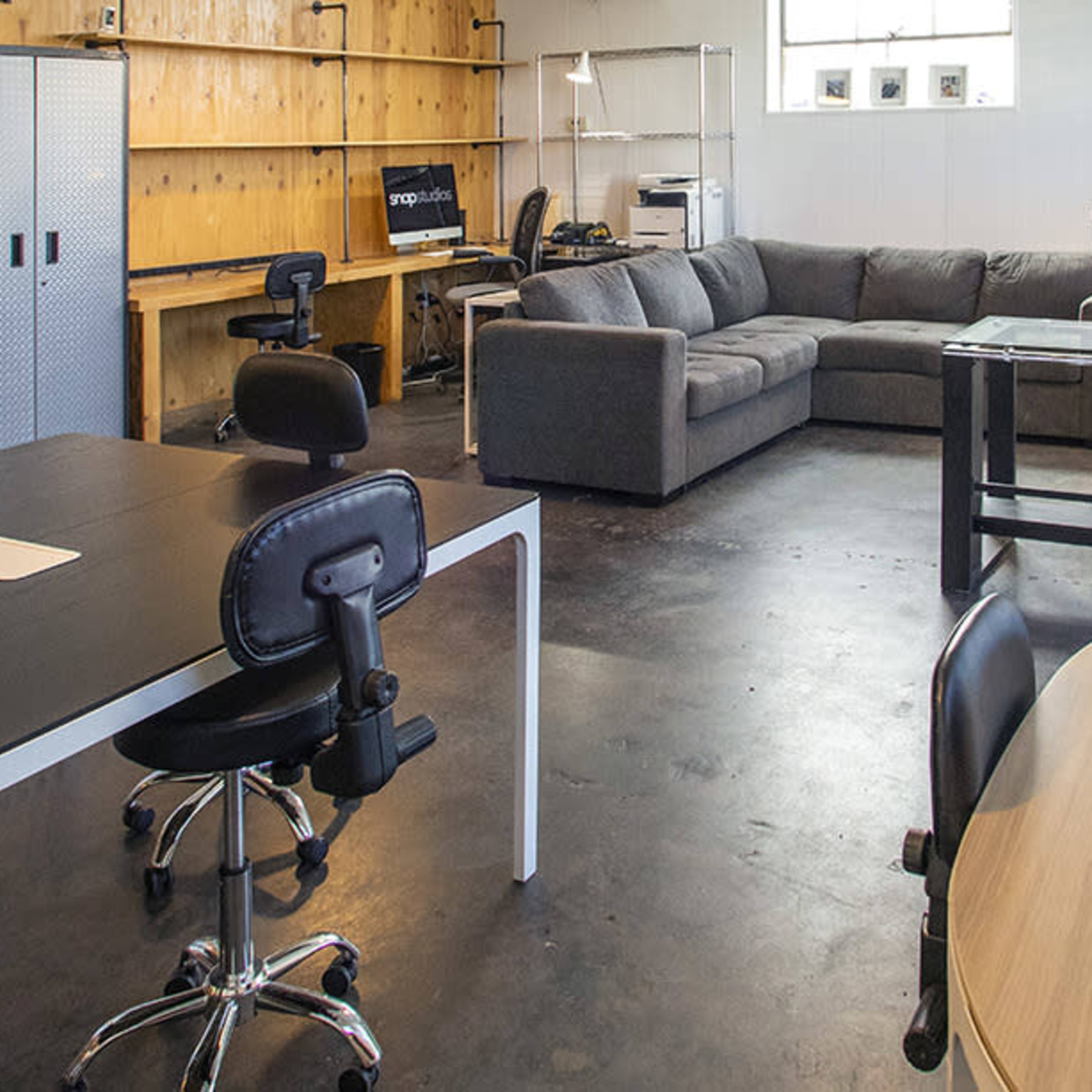 A modern office space featuring a large meeting table with rolling chairs, a cozy seating area with a sectional couch, and various workstations with computers.