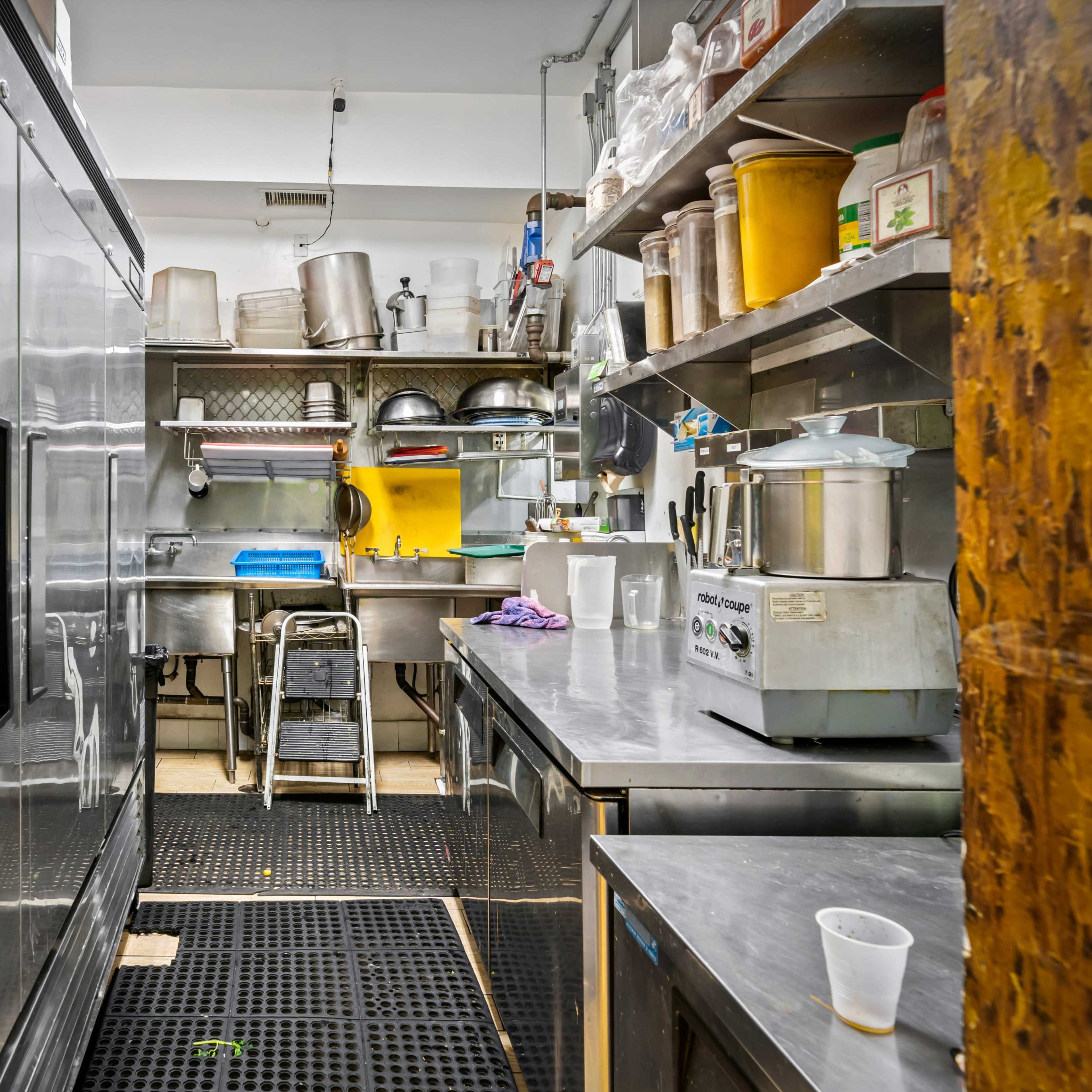 A narrow kitchen with stainless steel equipment, shelves filled with utensils and containers, and a side entrance to the cooking area.
