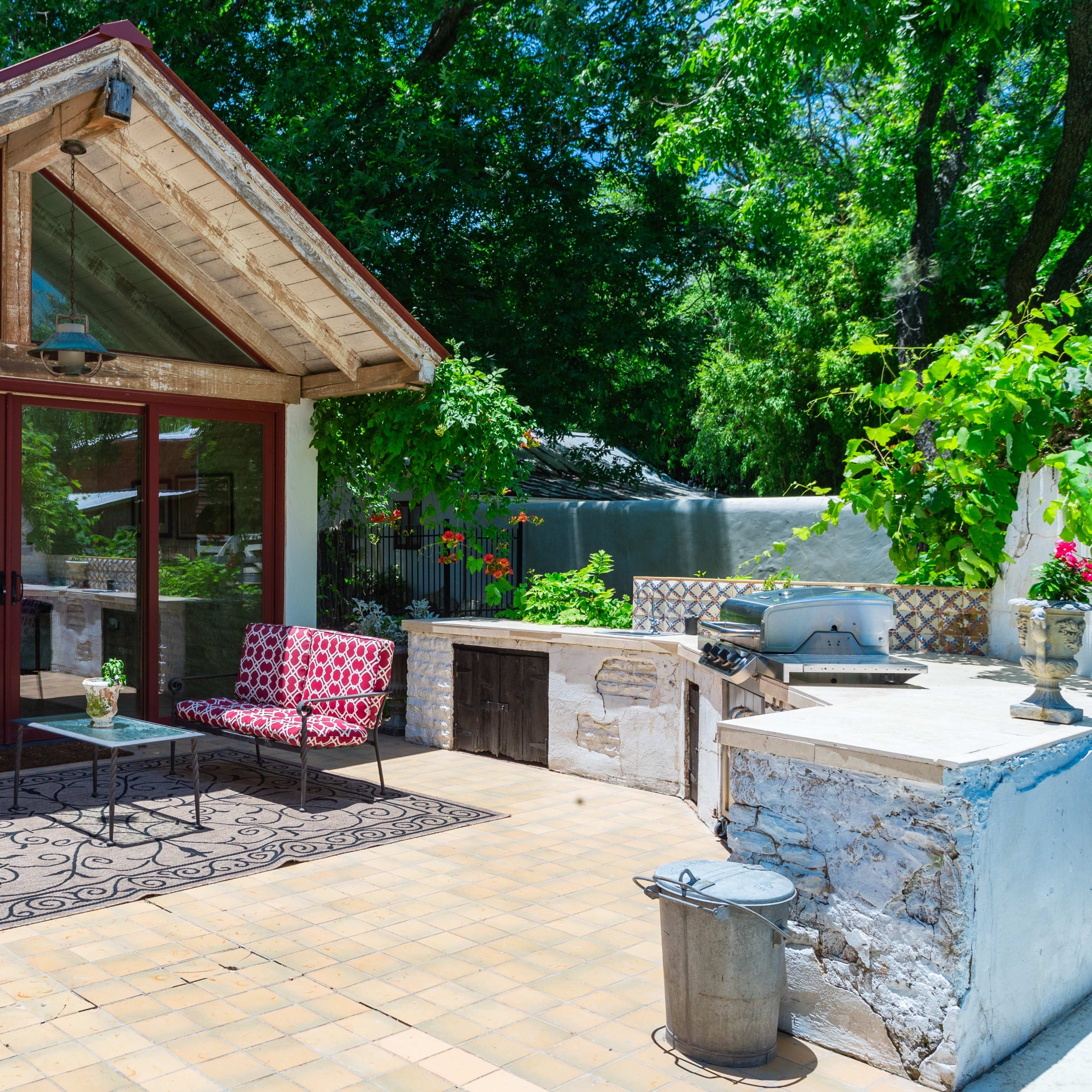 A patio area with two patterned chairs, a glass-fronted building, and a stone grill under a bright blue sky.