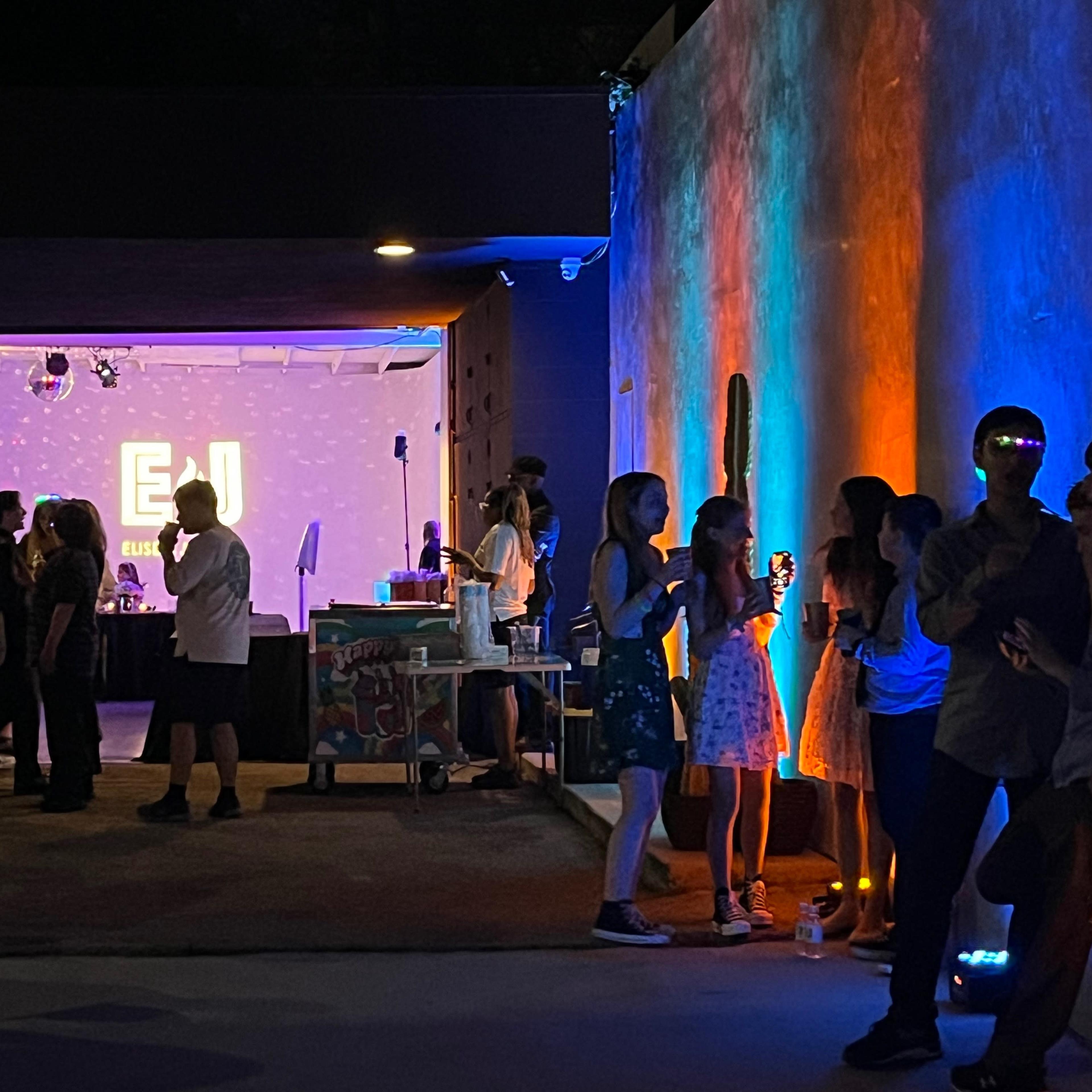 A lively event with people socializing under colorful lighting, with a projection displaying "EJ" in the background.