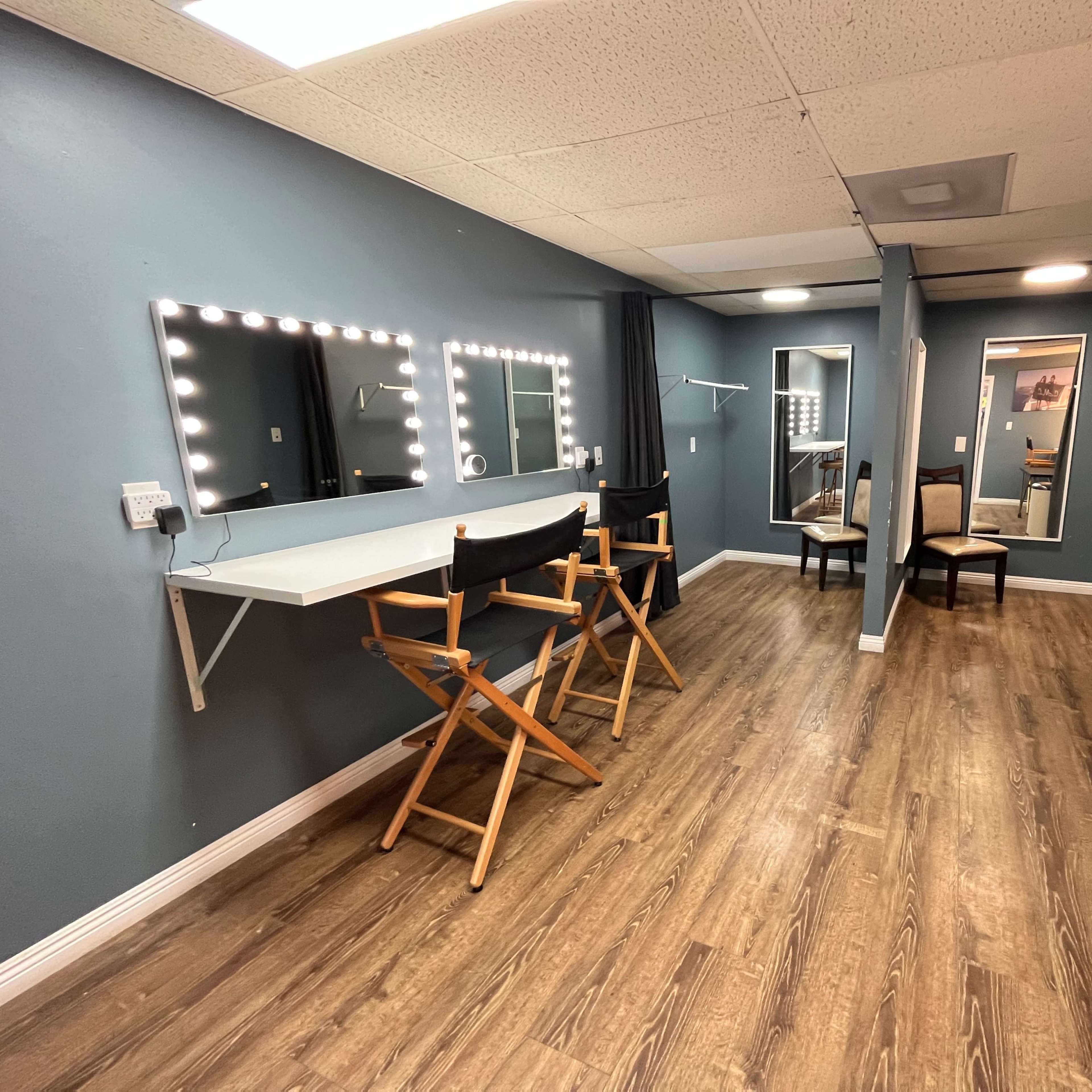 A well-lit dressing room with three makeup mirrors, two chairs, and a mirror at the far end.