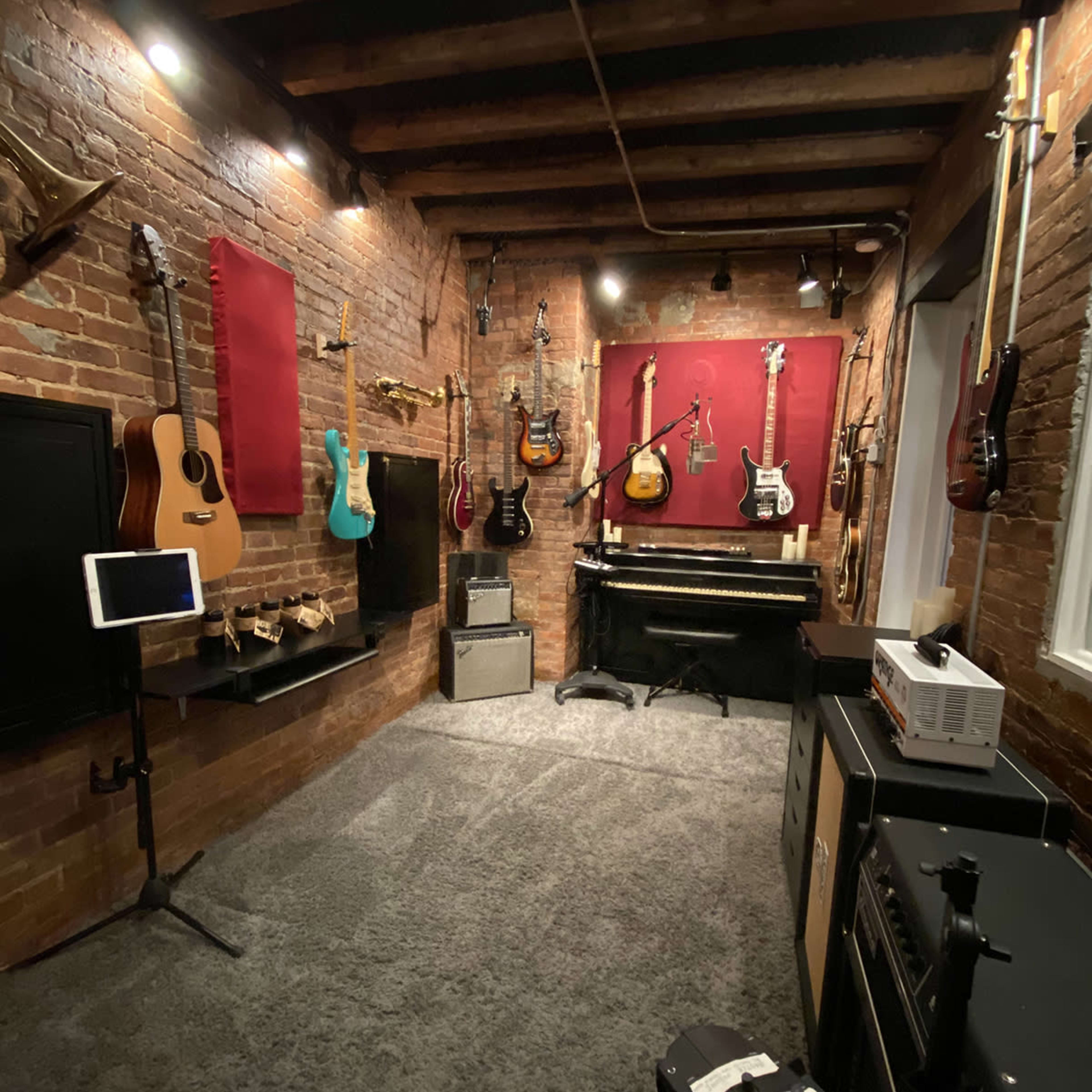 A music studio featuring an assortment of guitars hanging on brick walls, a piano in the corner, and various amplifiers and equipment on the floor.
