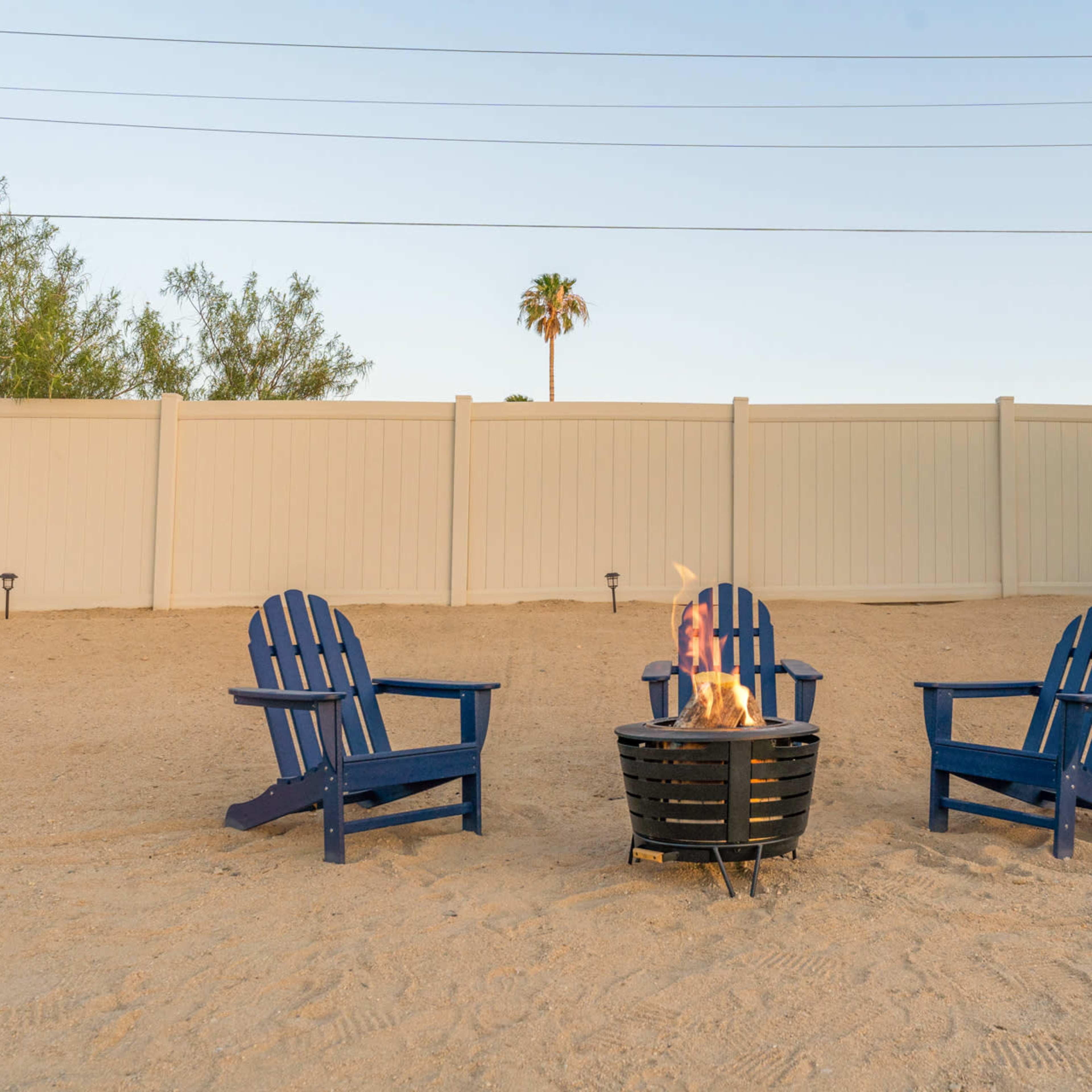 Three blue Adirondack chairs are arranged around a fire pit on a sandy area, with a palm tree visible in the background behind a white fence.